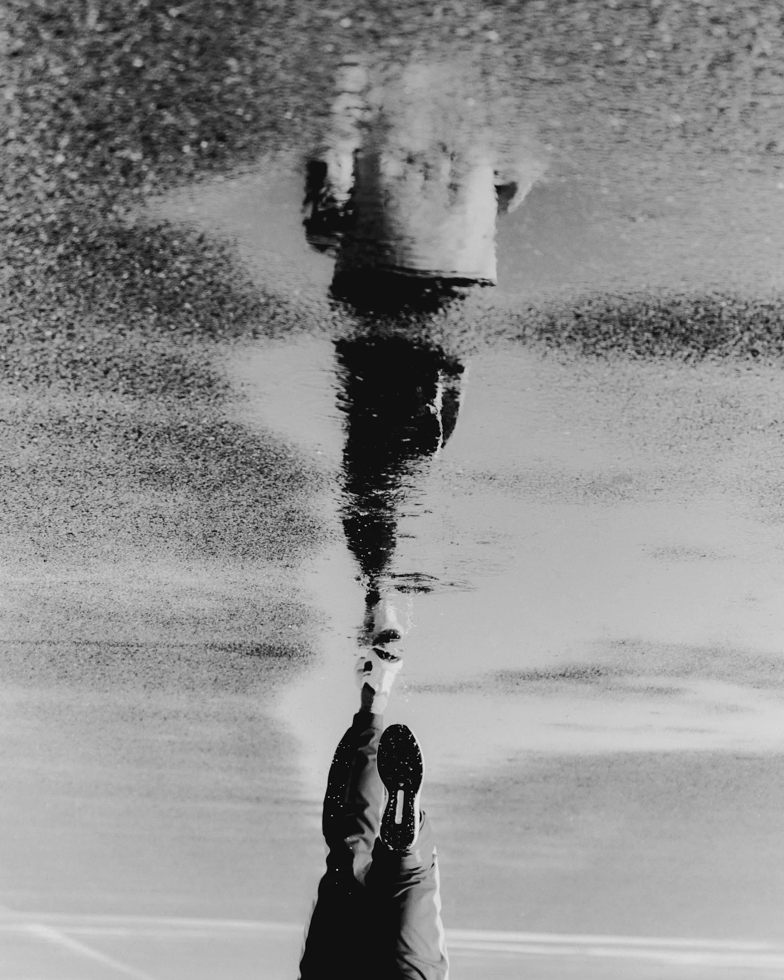 A person walking on a beach, reflected in a puddle, creating a mirrored effect of the person and the shoreline in the water in black and white.