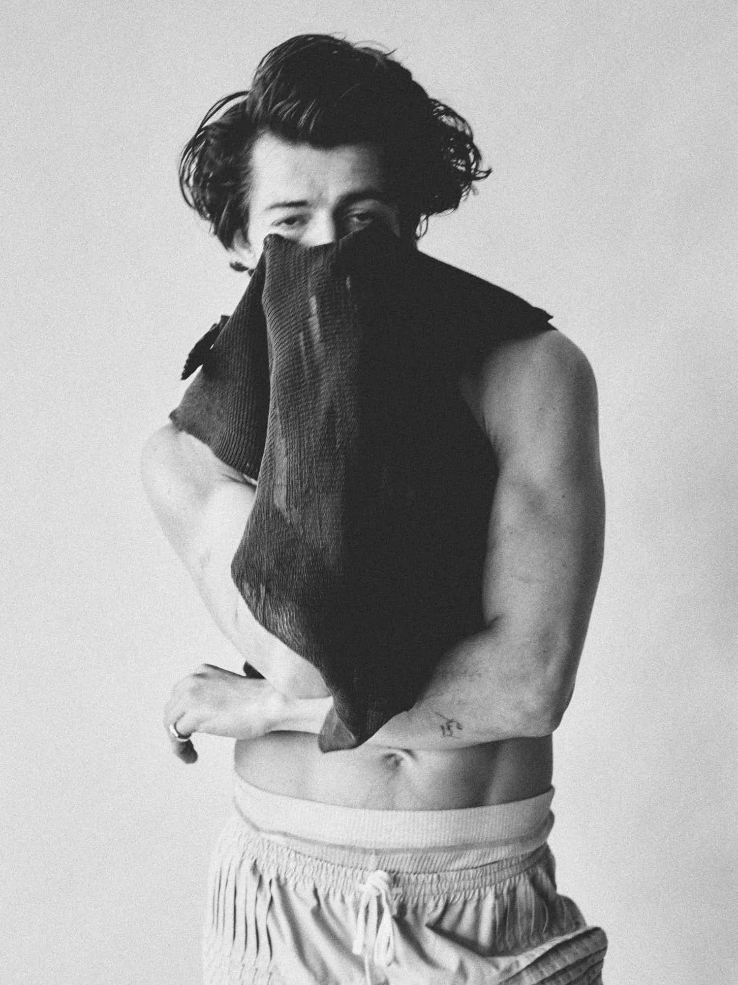 A black and white photo of a person with wavy hair covering part of their face, wearing a sleeveless top and baggy shorts, holding a piece of cloth or fabric in front of their face.