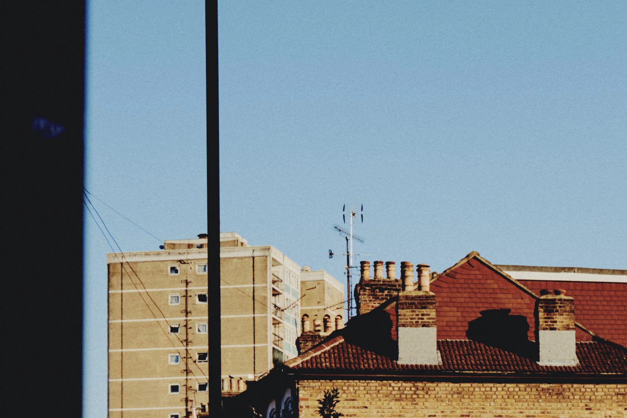 View of rooftops, a high-rise building, and rooftops with chimneys and TV antennas under a clear blue sky.