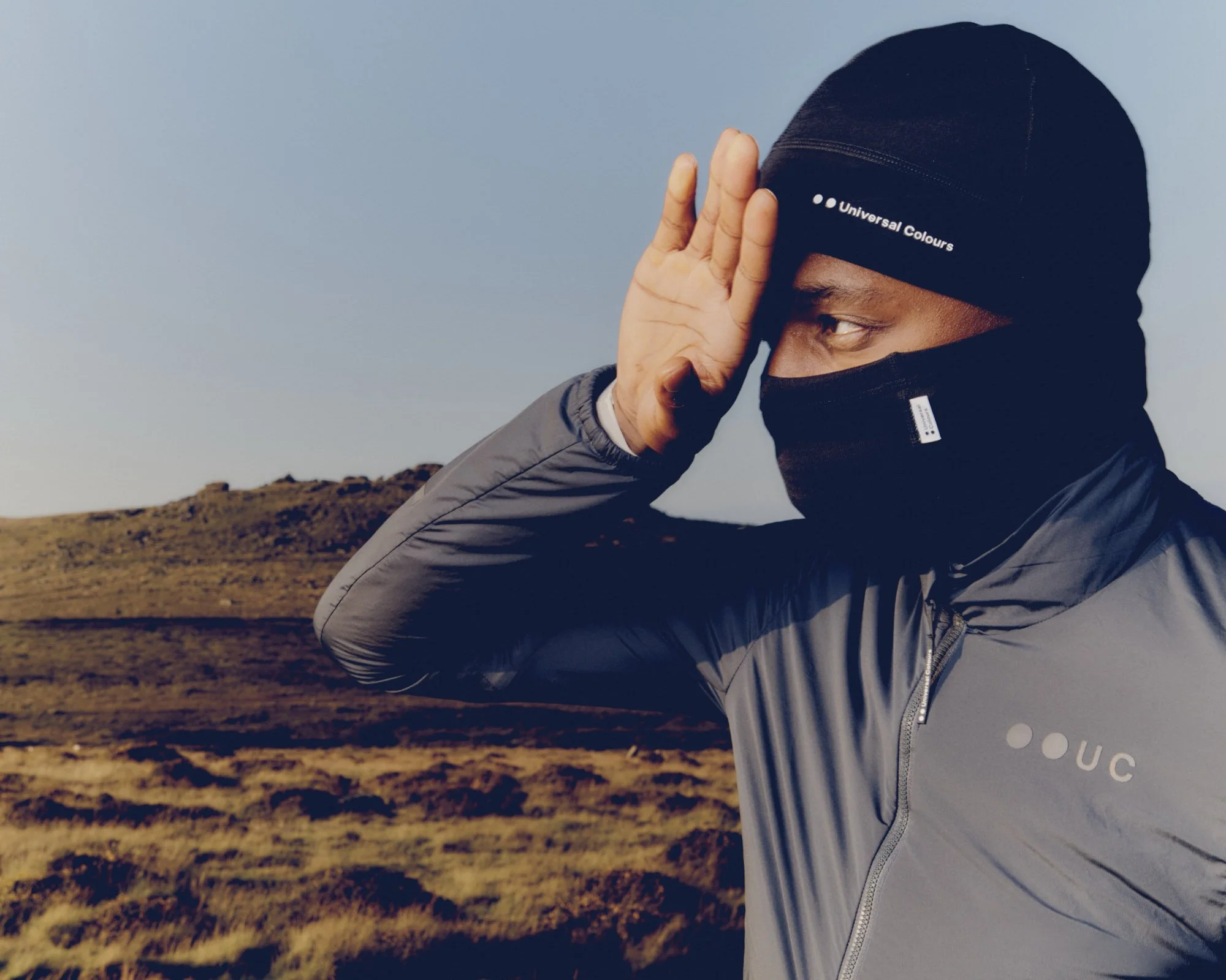 A person wearing a black beanie and a gray jacket, standing outdoors in a barren landscape under a clear sky, saluting with their right hand.