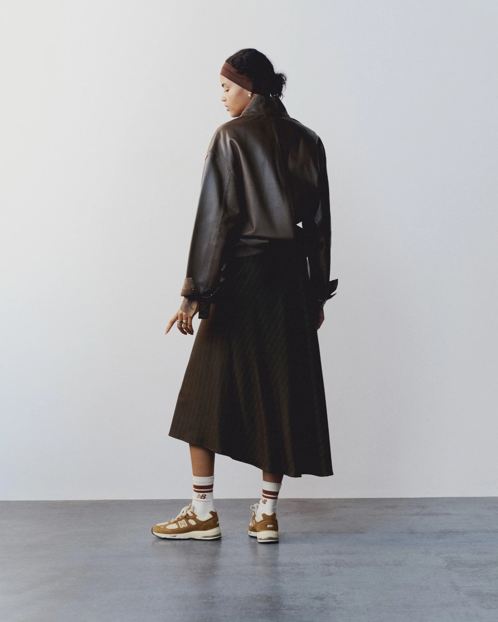 A woman standing against a plain white background, wearing a brown leather jacket, a dark green skirt, white socks with brown and white sneakers, a brown headband, and earrings, with her eyes closed and her head slightly tilted down.