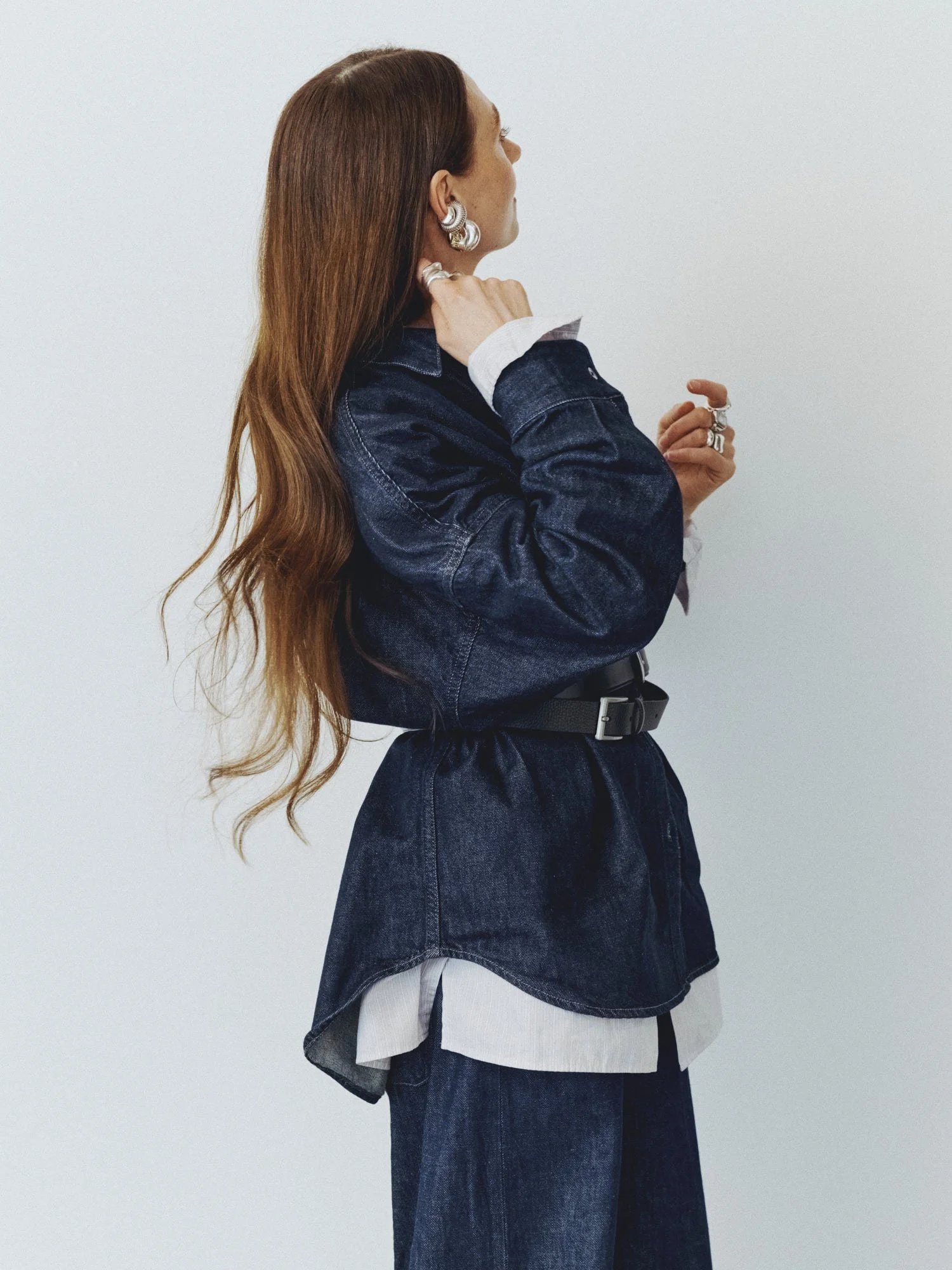 Side view of a woman with long, wavy auburn hair, wearing jewelry and a denim jacket, standing against a plain white background.