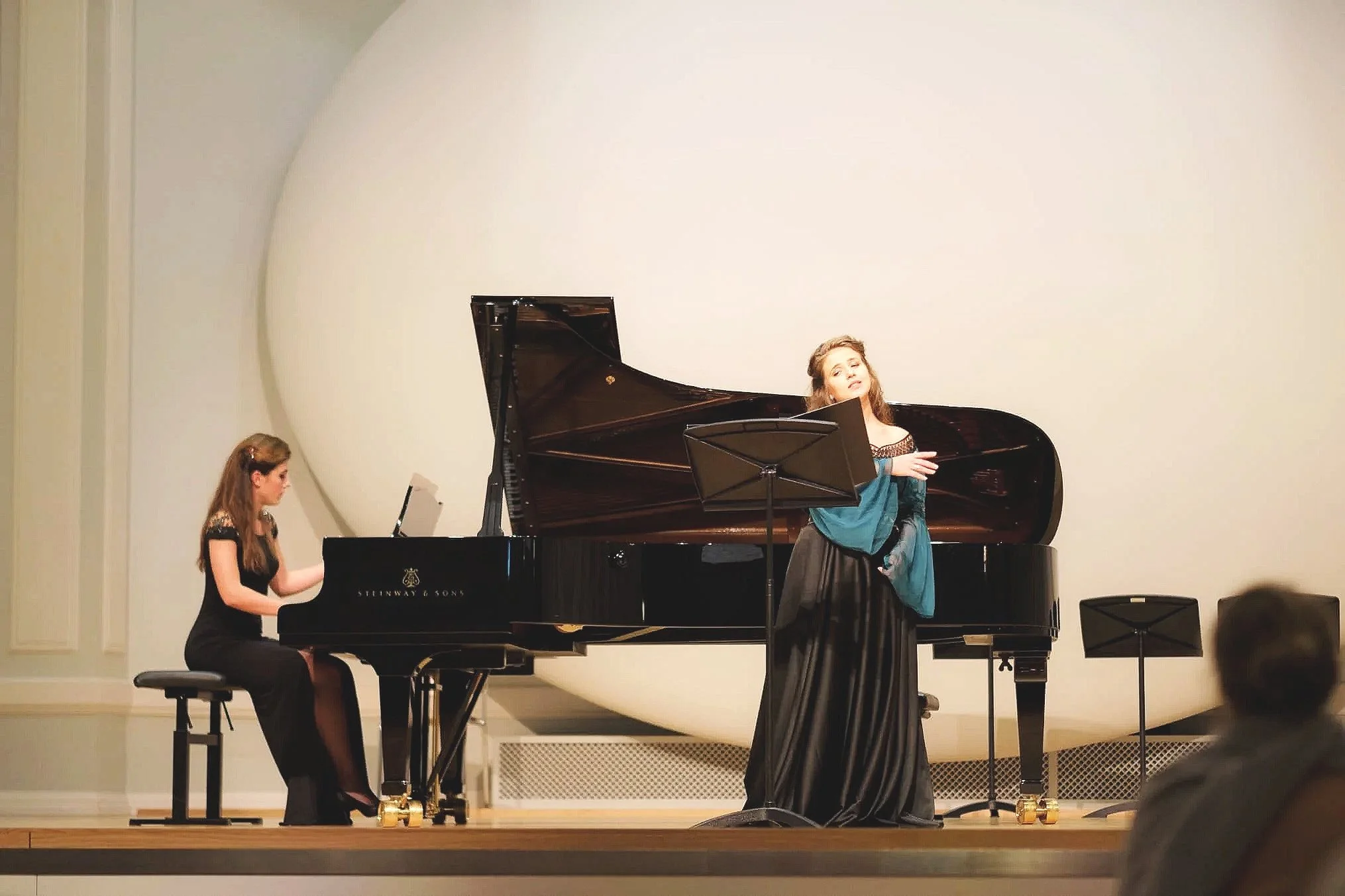 Two young women are performing a piano and a vocal solo on stage in a concert hall. The woman on the left is playing a grand piano, and the woman on the right is standing in front of a music stand, singing.