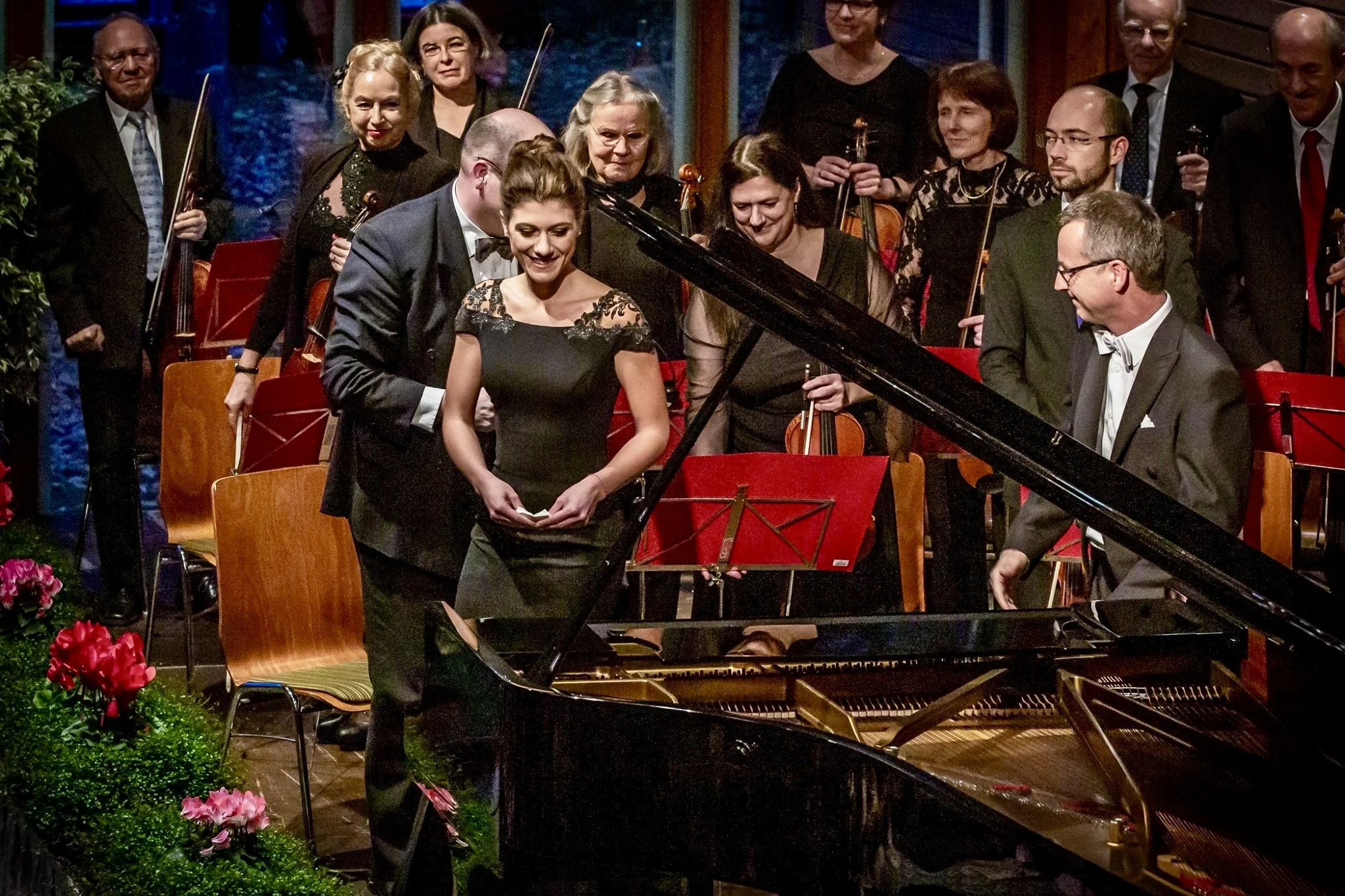 An orchestra performing on stage, with a young woman in a black dress at a grand piano in the foreground. Musicians are holding violins and other string instruments, and some are holding drinks. The setting appears to be a formal concert or event, with decorative plants and a warm ambiance.