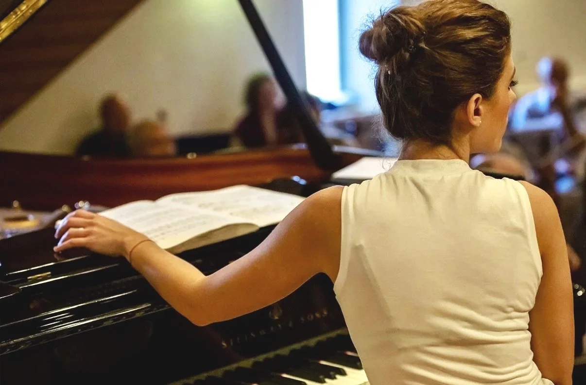 A woman with her hair styled in a bun playing a grand piano in a room with an audience.