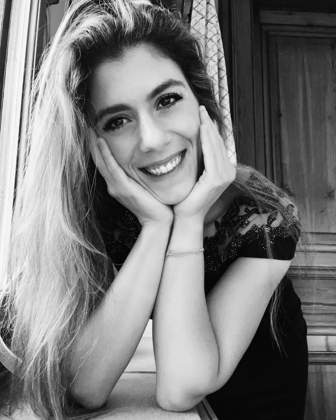 A young woman with long hair smiling and resting her face in her hands, in black and white.