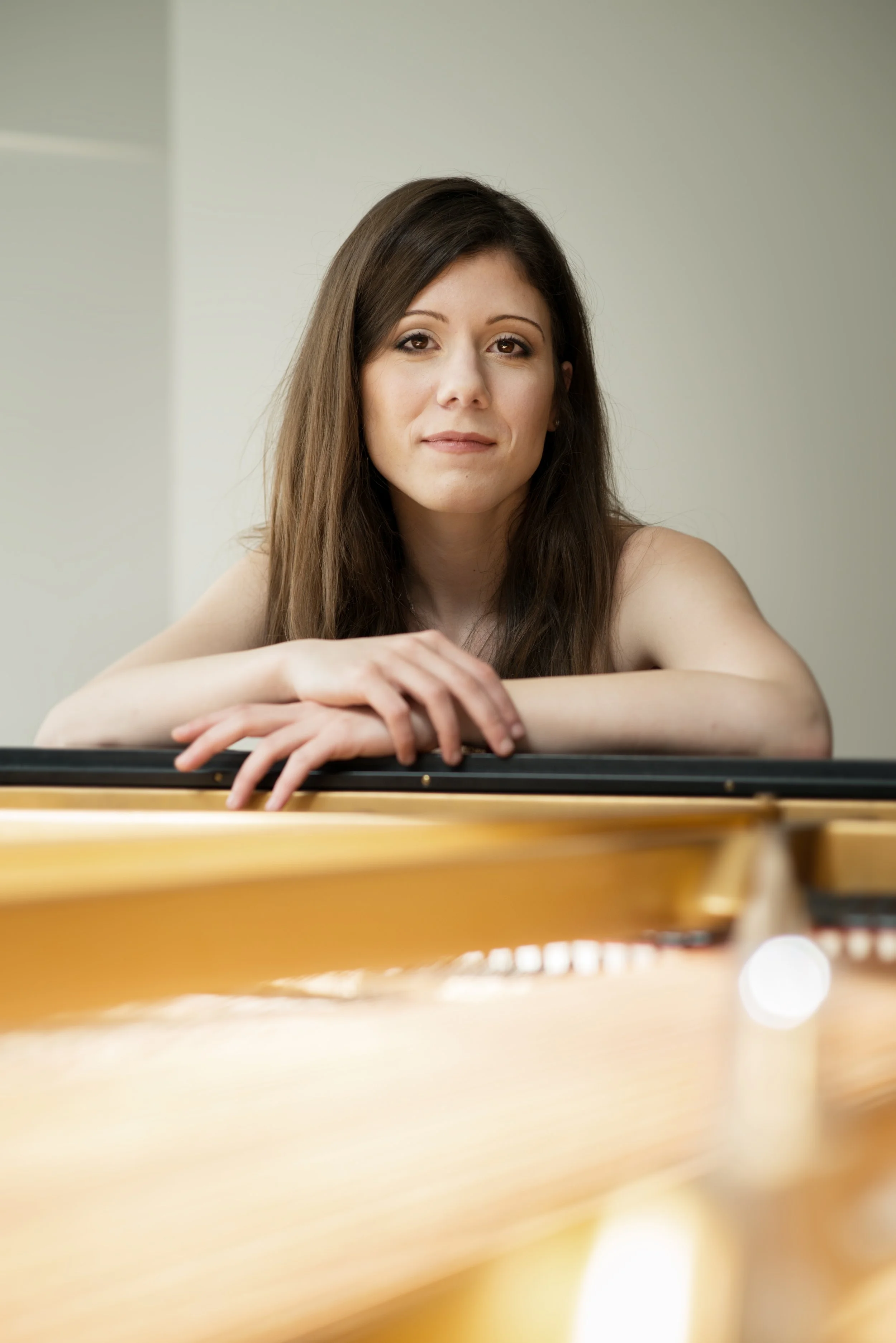 A woman with long brown hair resting her arms on a piano, looking at the camera with a slight smile, in a room with plain light-colored walls.