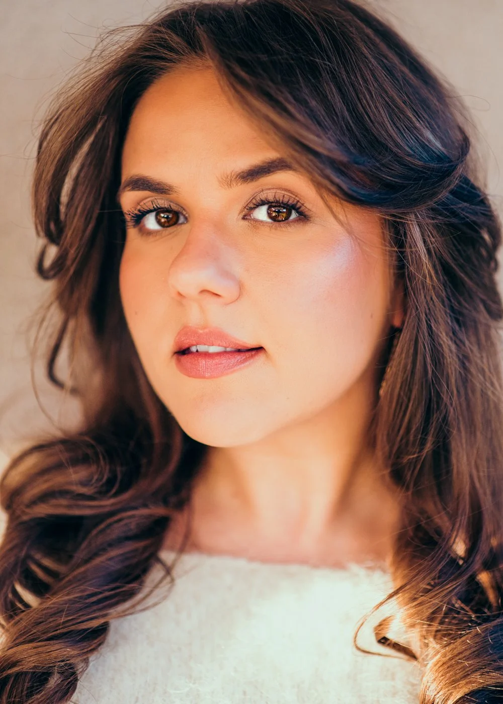 Close-up portrait of a young woman with long, wavy brown hair and brown eyes, wearing natural makeup and a light-colored top.