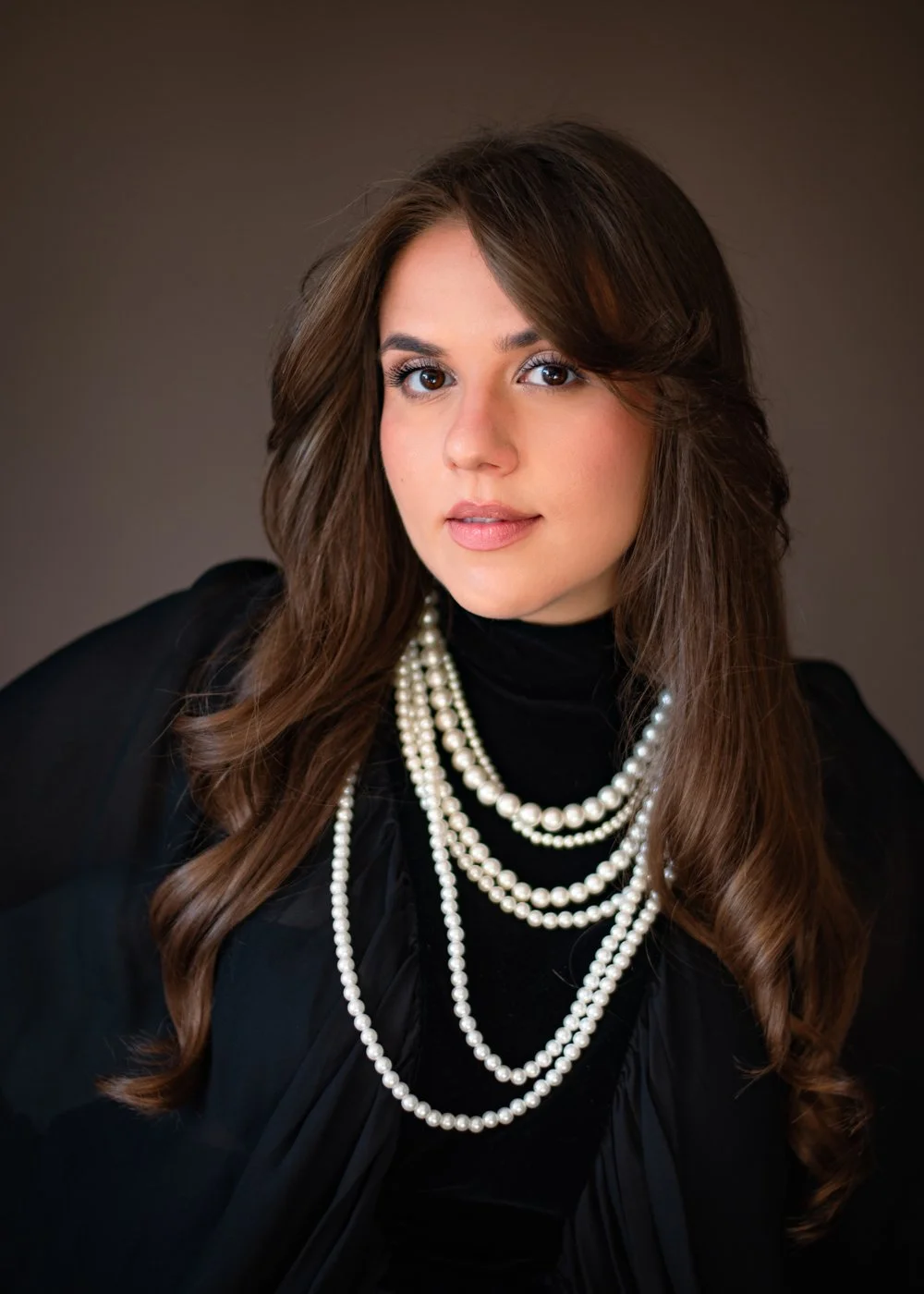 A woman with long, wavy brown hair, wearing a black top and multiple strands of white pearls, posing against a neutral background.