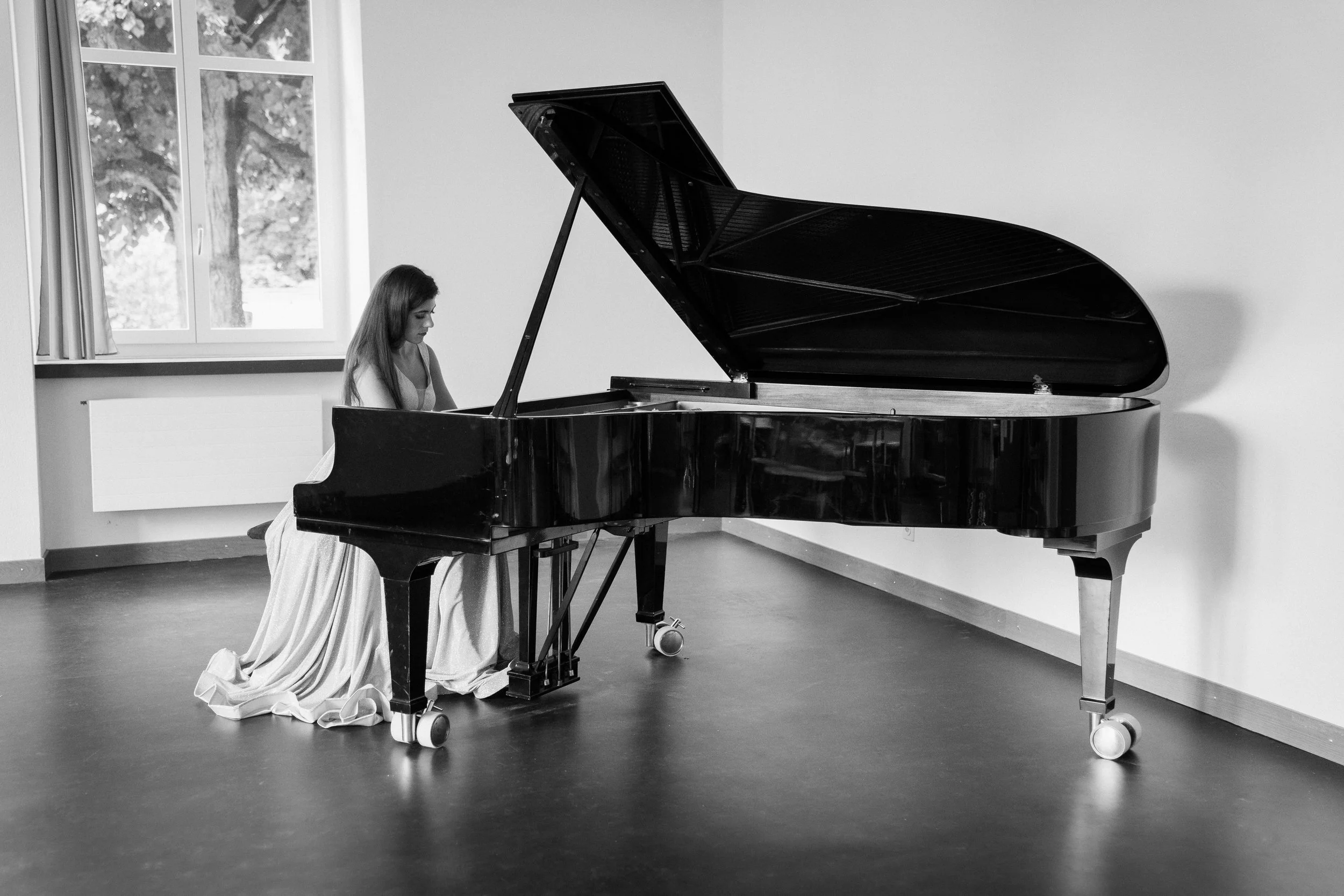 A young girl in a long dress playing the piano in a room with a large window showing trees outside.