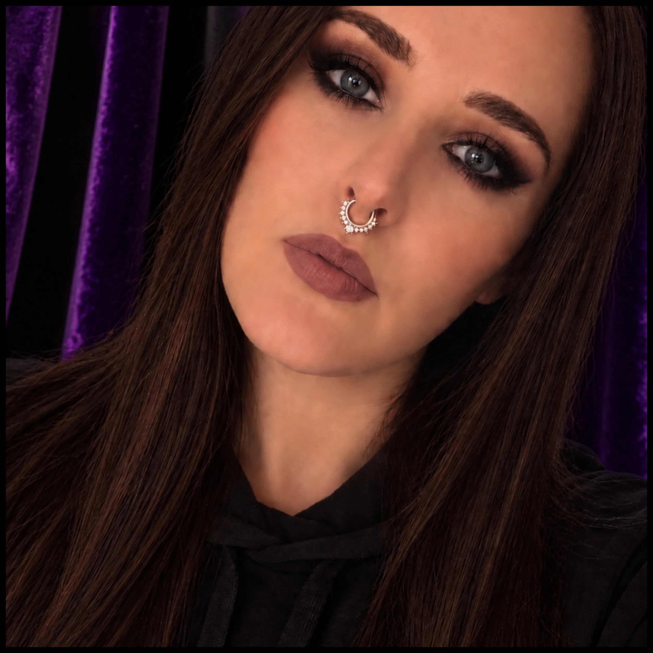 A woman with long brown hair, wearing a black headscarf, black lipstick, and dramatic eye makeup, against a purple textured background.