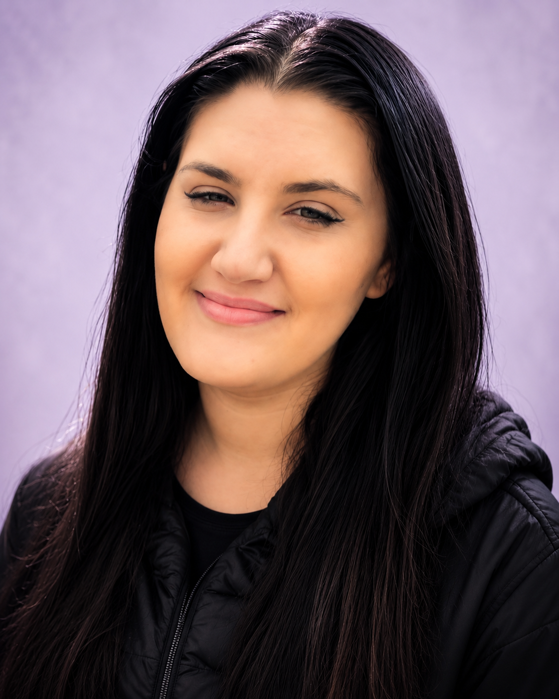 Close-up portrait of a young woman with long black hair, smiling slightly with one eye squinting, wearing a black jacket, against a purple background.