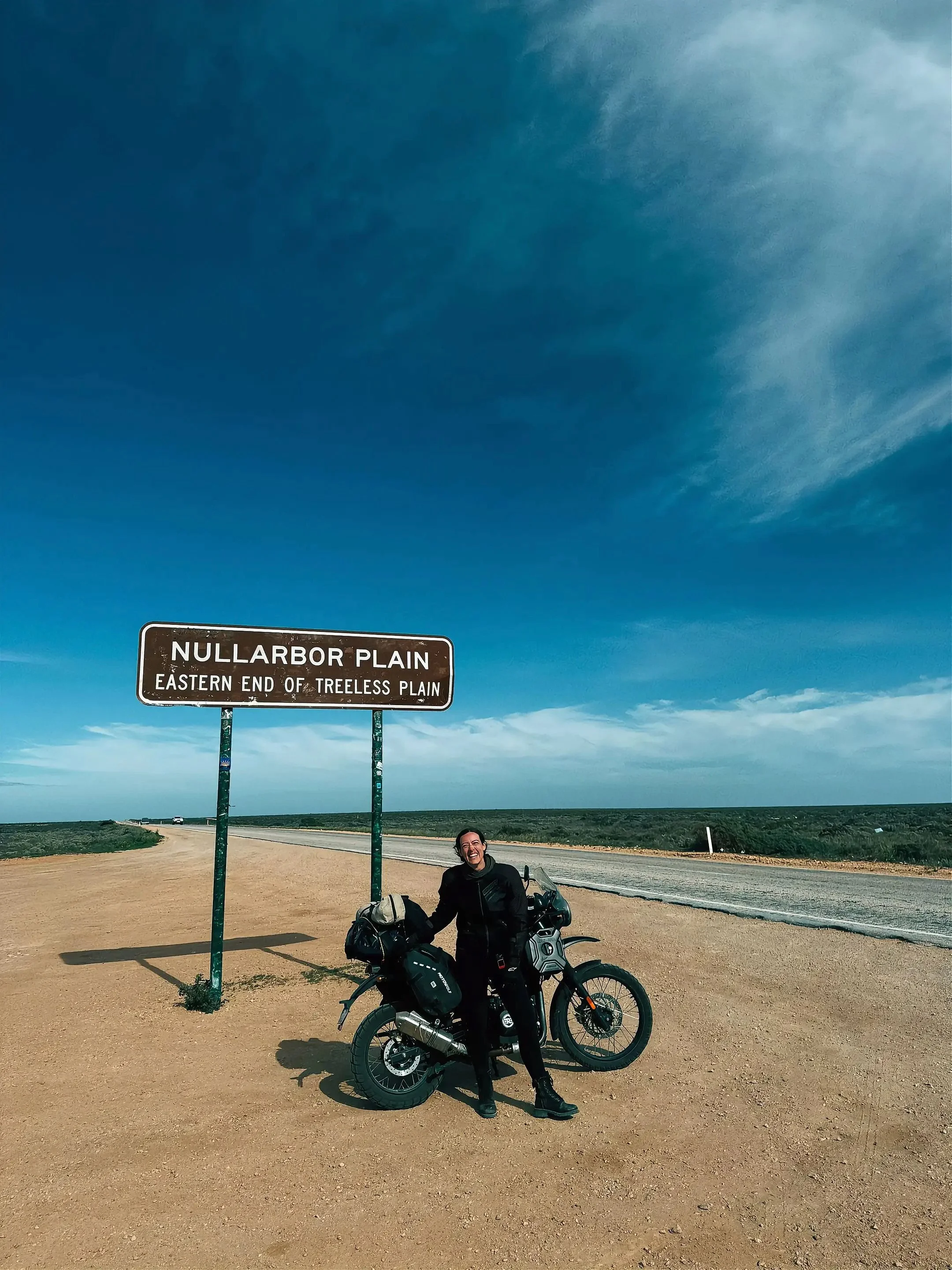 A smiling woman standing next to a motorcycle with loaded saddlebags, in a rural area under a blue sky, next to a road sign that reads Nullarbor Plain, Eastern End of Treeless Plain.