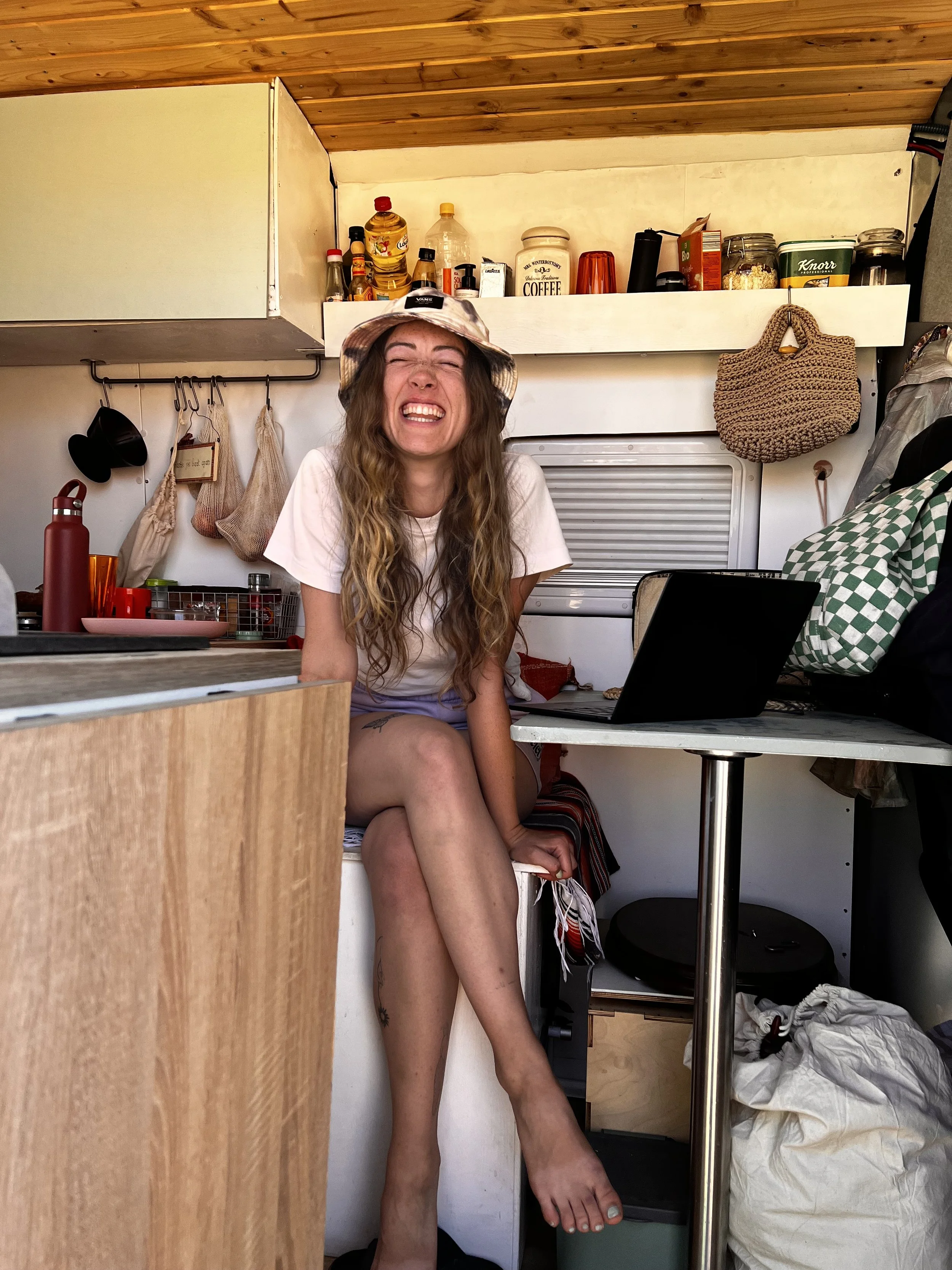 A woman with long wavy hair wearing a bucket hat smiling broadly with closed eyes, sitting inside a small kitchen space.