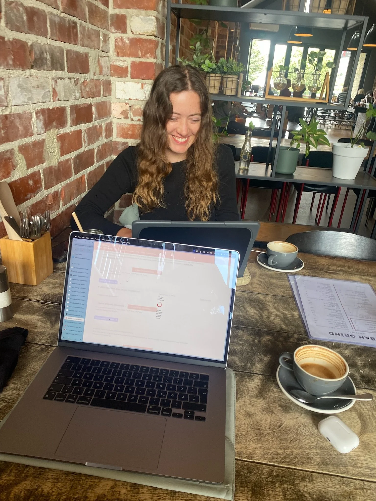 A woman with long wavy hair smiling while sitting at a table in a cozy café. There are two cups of coffee on the table, a laptop, and a menu. The background has a brick wall and plants.
