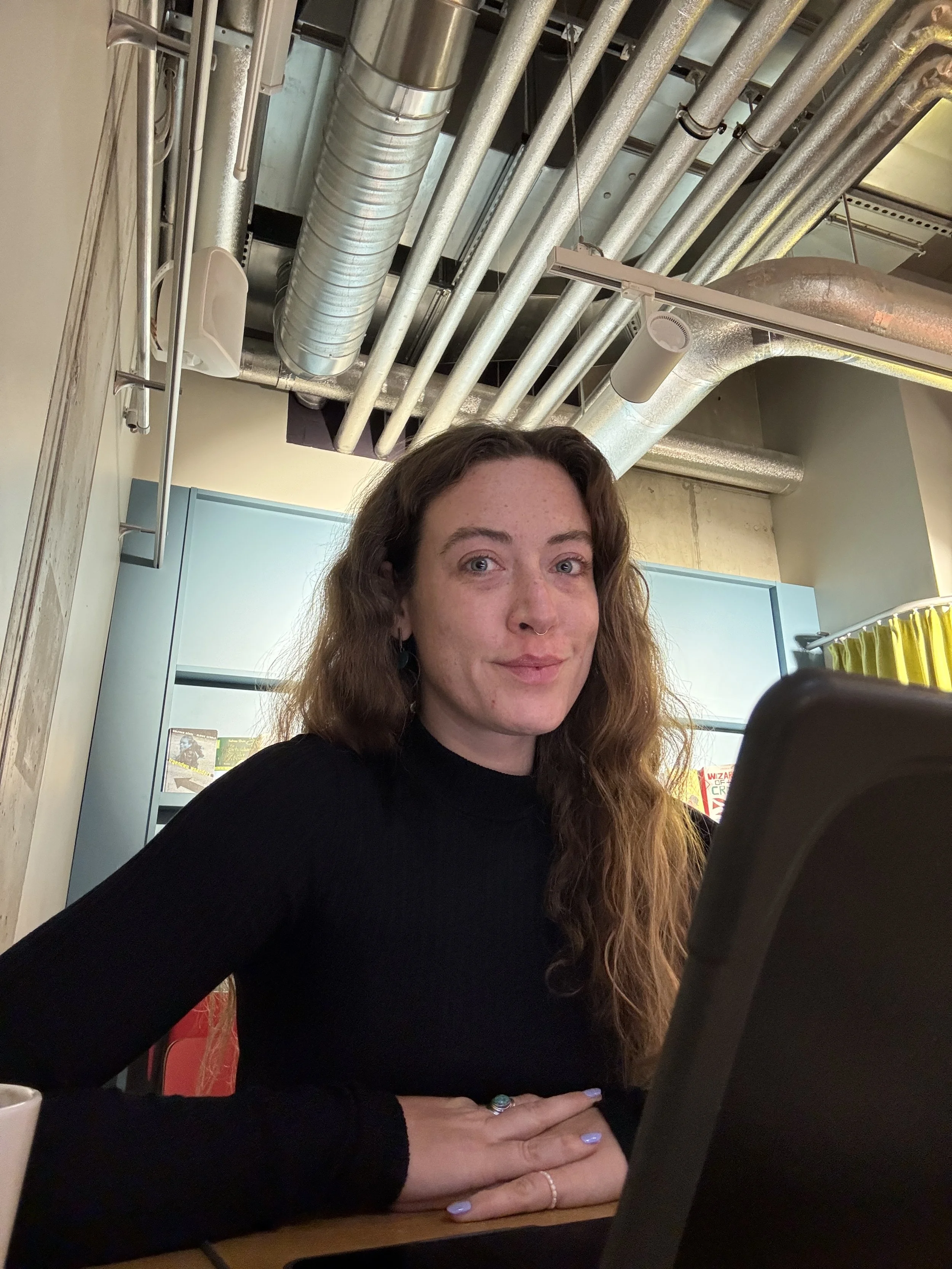 A woman with long, curly hair, wearing a black long sleeve shirt, sitting at a table and looking at a device. The background includes aired ductwork, a blue cabinet, and a yellow curtain.