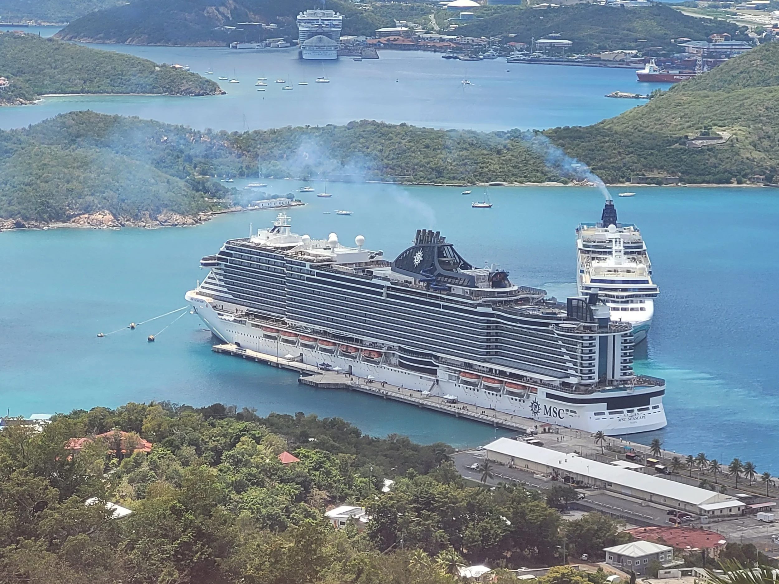 Two large cruise ships docked at a harbor surrounded by lush green hills, with smaller boats in the water and a scenic landscape of islands and a coastal town.