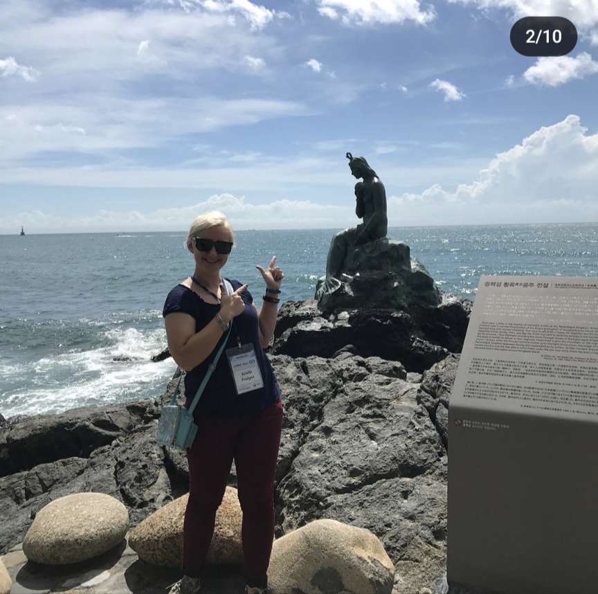 A woman photobombing a seaside sculpture of a mermaid with the ocean and sky in the background.
