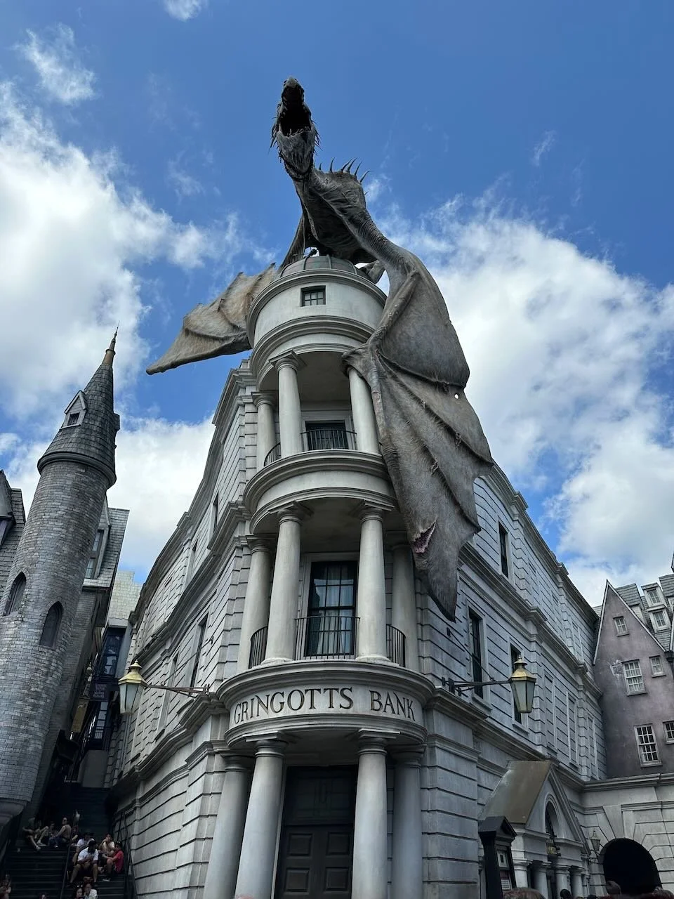 A large dragon statue is perched on top of Gringotts Bank building found in Universals Diagon Alley, with the dragon's mouth open, set against a partly cloudy sky.