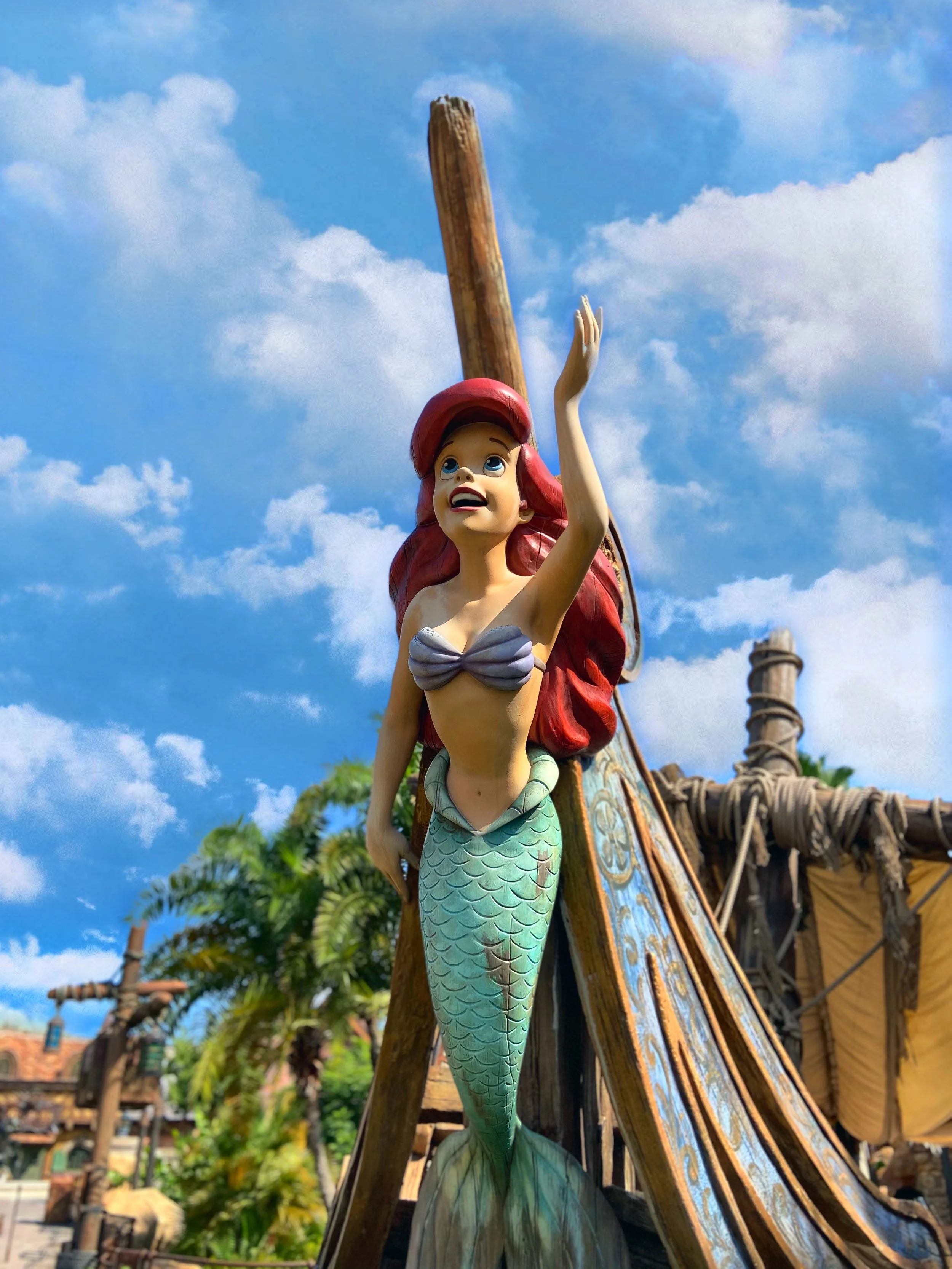 Colorful Ariel the little mermaid sculpture with red hair and blue eyes, holding a wooden mast, on a pirate ship, under a blue sky with white clouds.