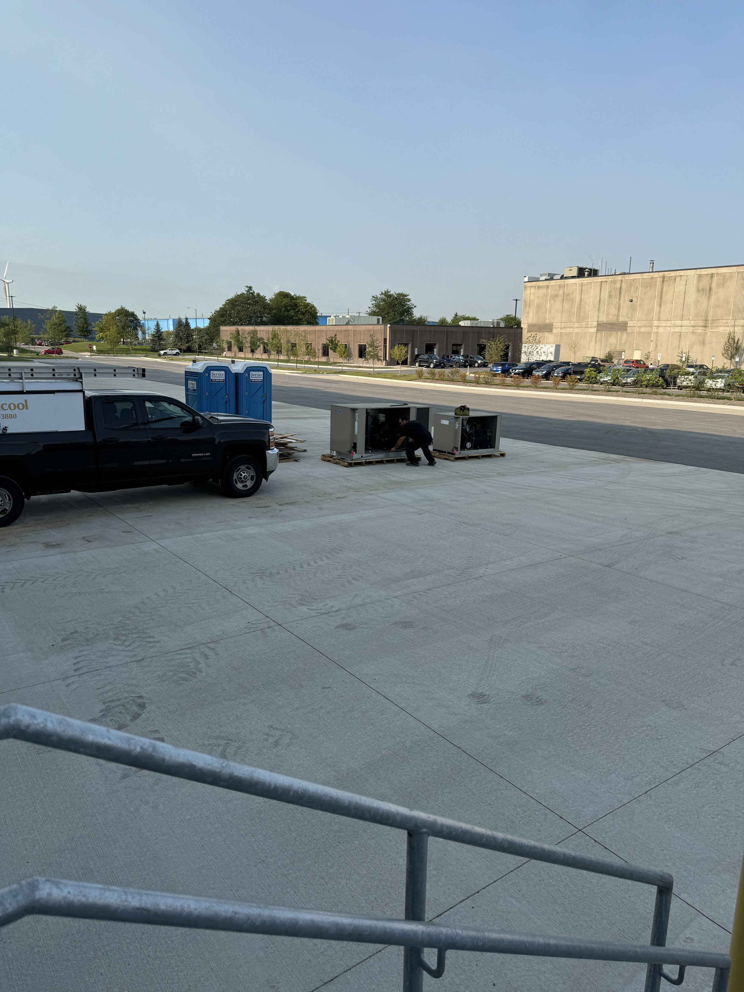 A parking lot with a black pickup truck, blue portable restrooms, and two large gray equipment units, with a worker crouching near one of the units. In the background, there are buildings, trees, and parked cars.