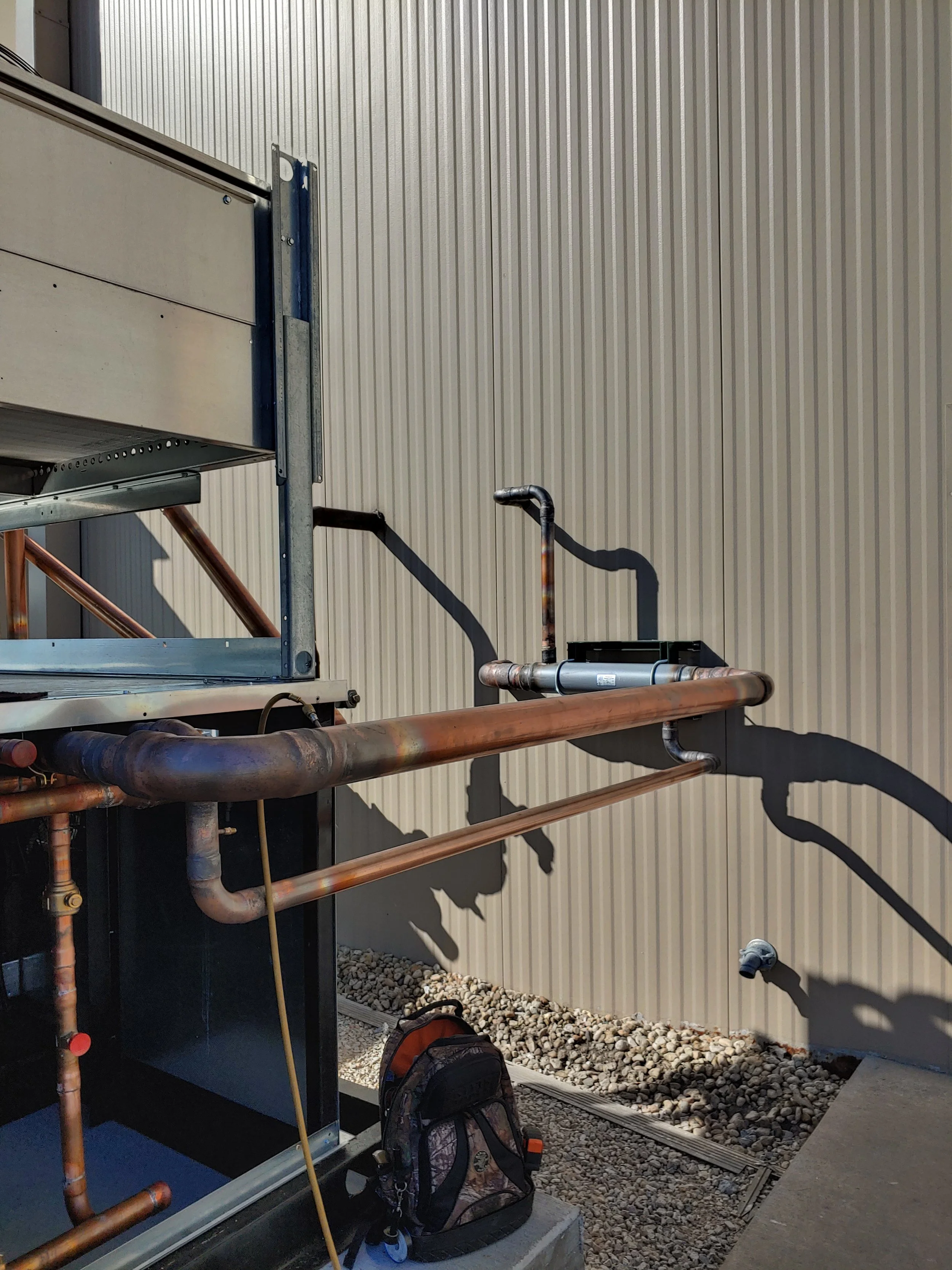 Side view of an industrial or commercial building exterior wall made of metal siding. There are visible copper pipes, vent pipes, and an HVAC unit. Two backpacks are on the ground, and shadows of pipes are cast on the wall.