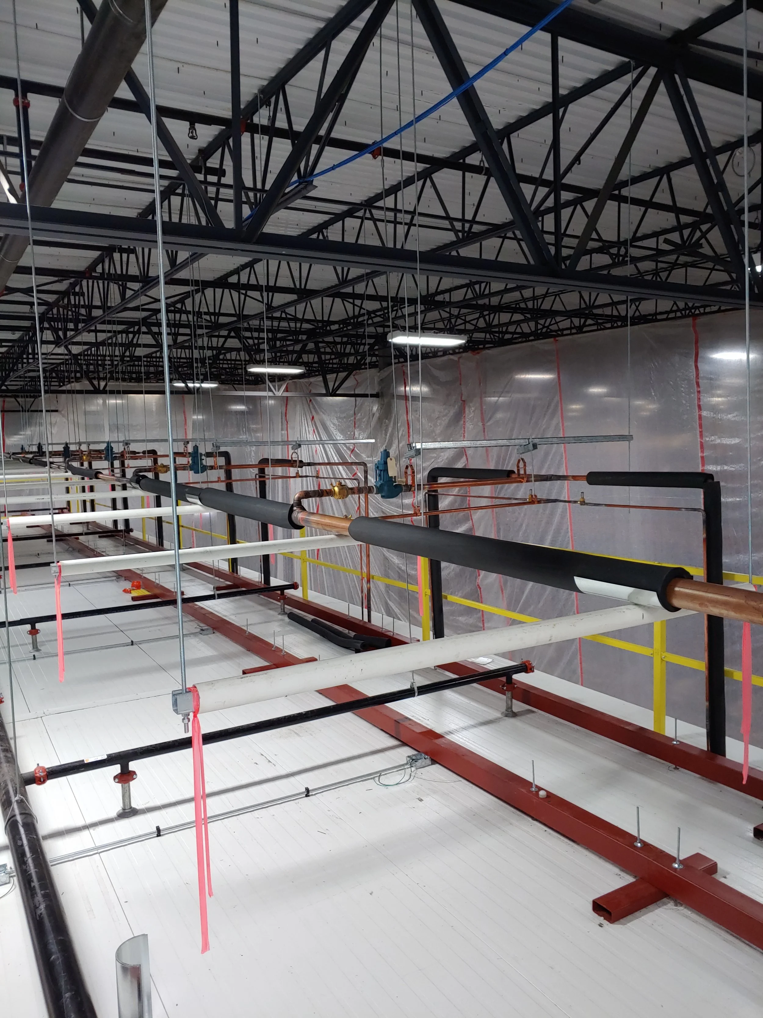 The image shows an industrial or construction setting with various pipes, metal beams, and scaffolding. The ceiling is constructed with a metal grid, and there are plastic sheeting and electrical wiring visible in the background.