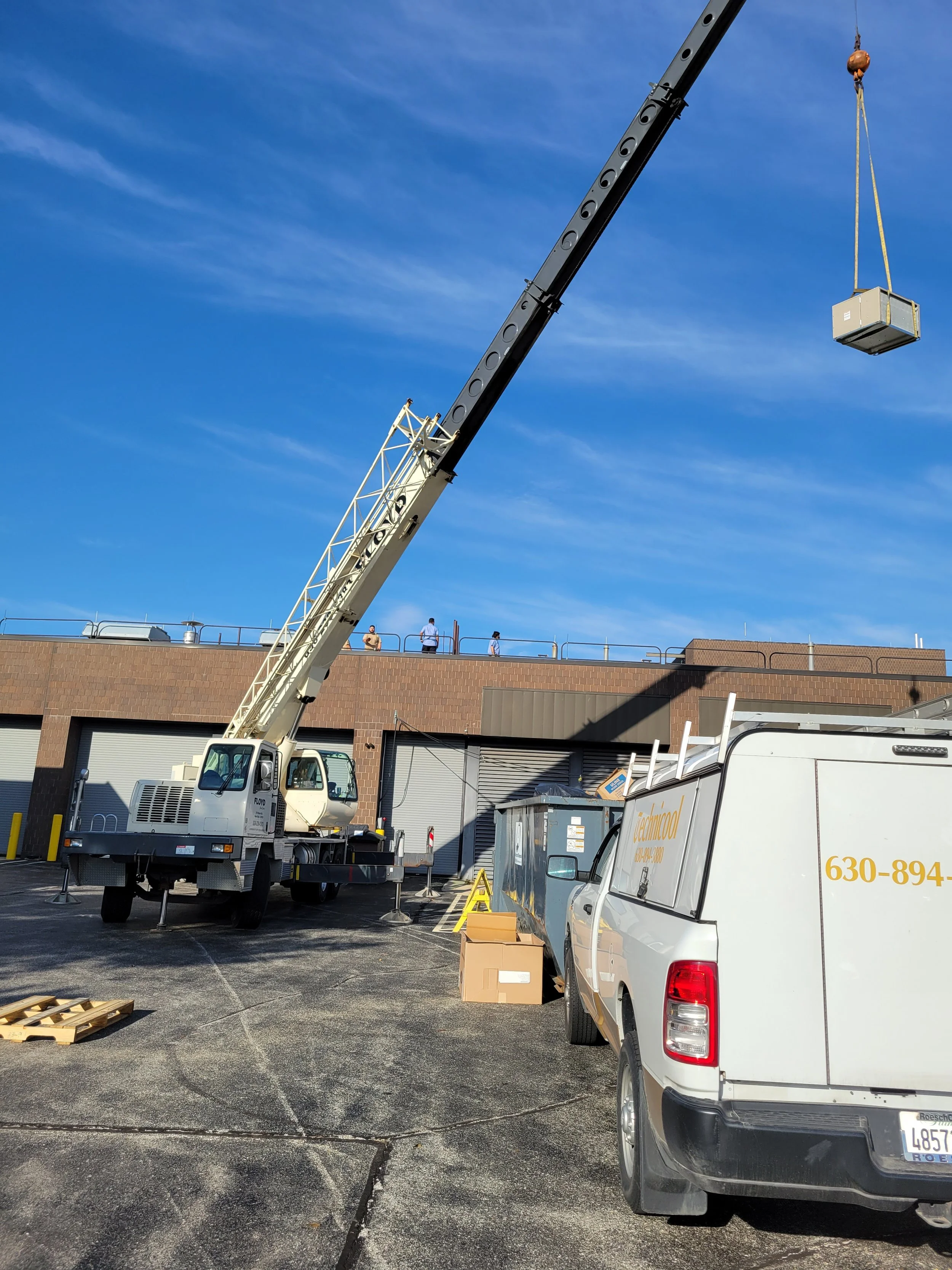 A crane lifting a large crate in a parking lot with a building in the background and a white van in the foreground.