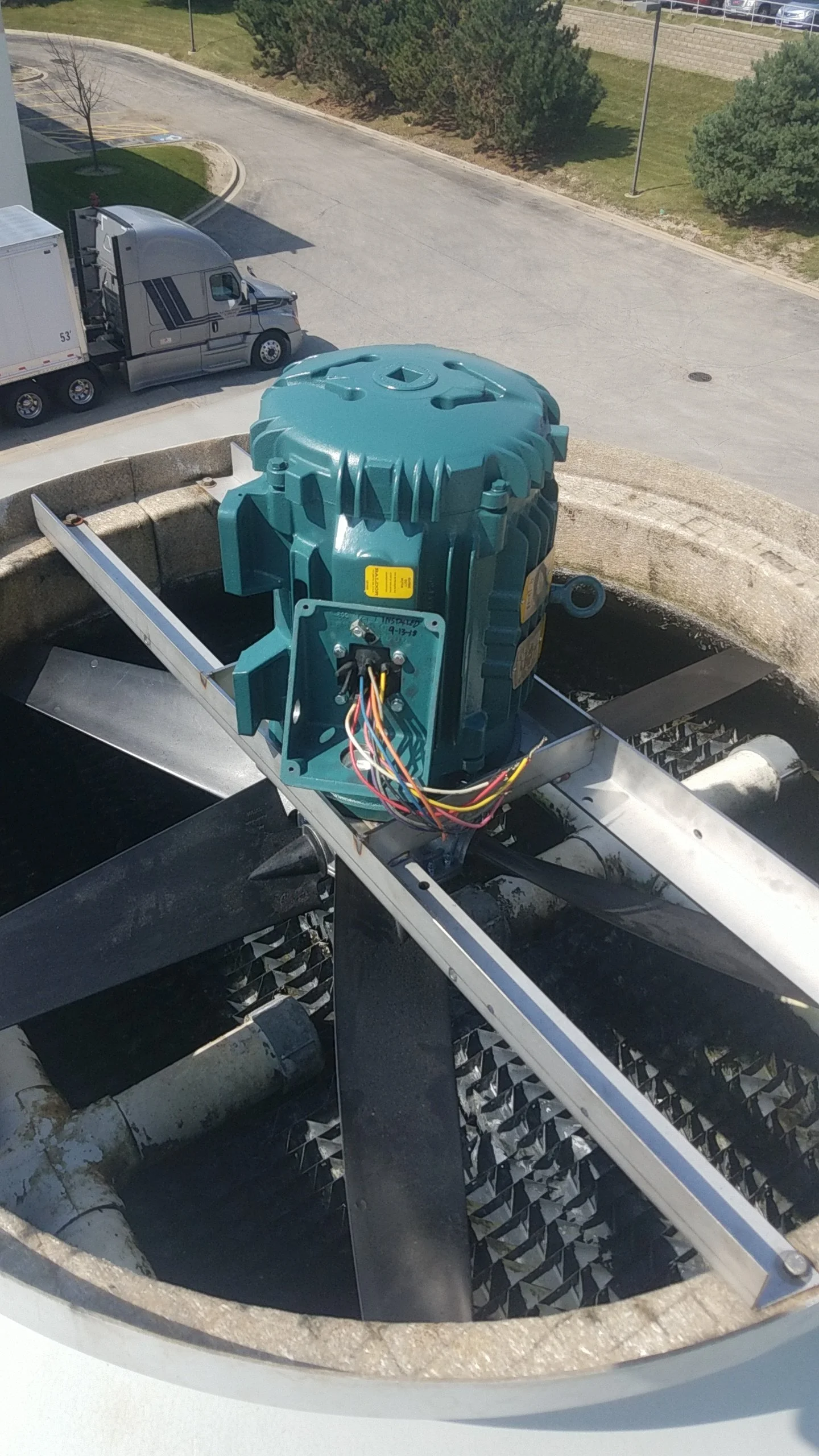 Close-up view of the internal components of a large industrial fan or blower, showing a motor with wires, mounted inside a metal housing with blades visible at the bottom, located outdoors near a parking lot.