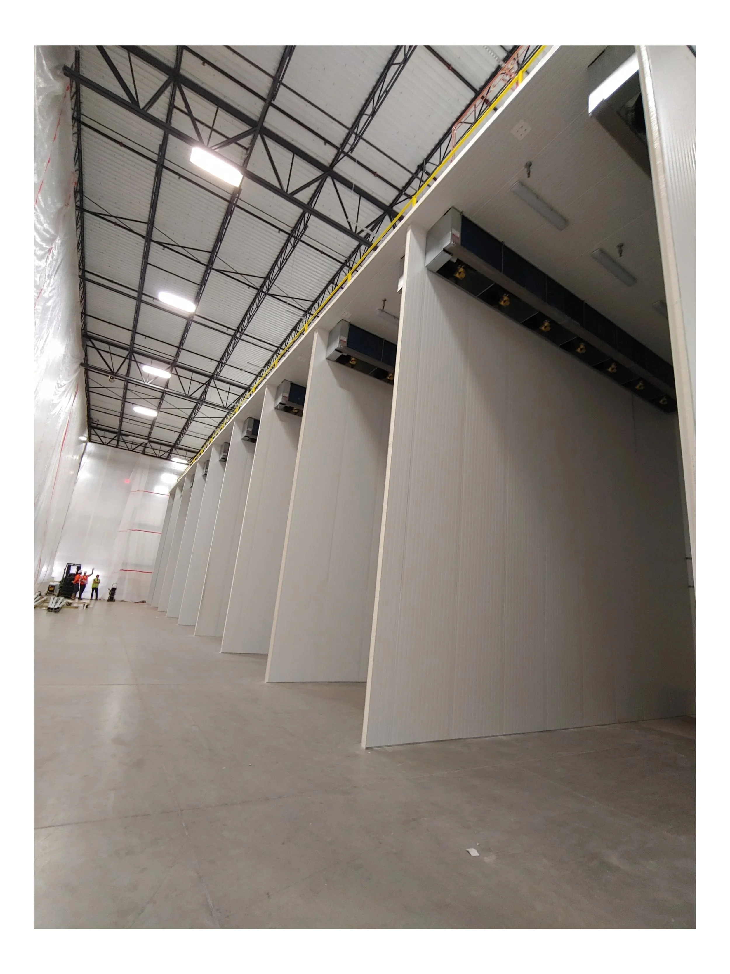 Large industrial warehouse with white partition walls, high ceiling with exposed steel beams, and workers in the distance.