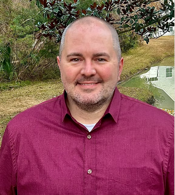 A smiling man with a beard and short hair standing outdoors near a body of water, with trees and bushes in the background.