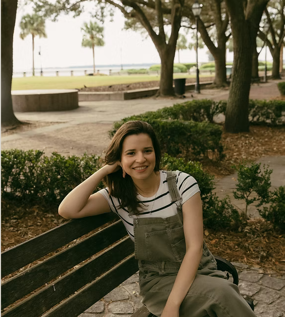 Young woman sitting on a park bench smiling, wearing a striped t-shirt and overalls, with trees and a waterfront in the background.