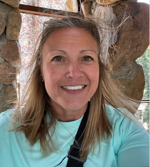 A woman with blonde hair smiling, wearing a light blue shirt, with rocky background and sunlight.