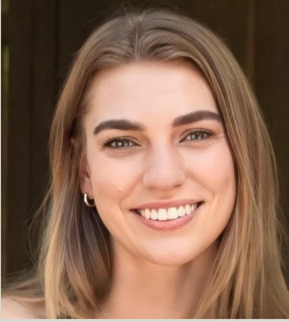 Close-up of a young woman with long blonde hair, smiling showing her teeth, wearing small hoop earrings, in front of a wooden background.