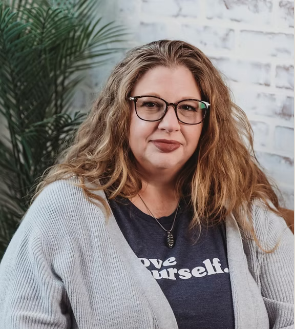 A woman with curly red hair and glasses, wearing a navy blue t-shirt with white text and a light gray cardigan, sitting indoors near a white brick wall and green plants.