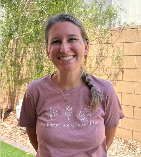 A woman smiling outdoors with a light purple t-shirt that has white floral illustrations and the phrase "the human spirit is resilient."