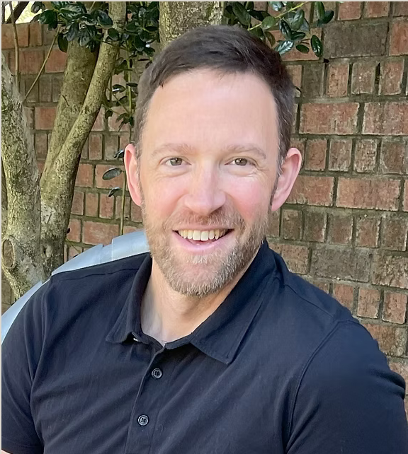 Man smiling outdoors in front of a brick wall and a tree, wearing a black polo shirt.