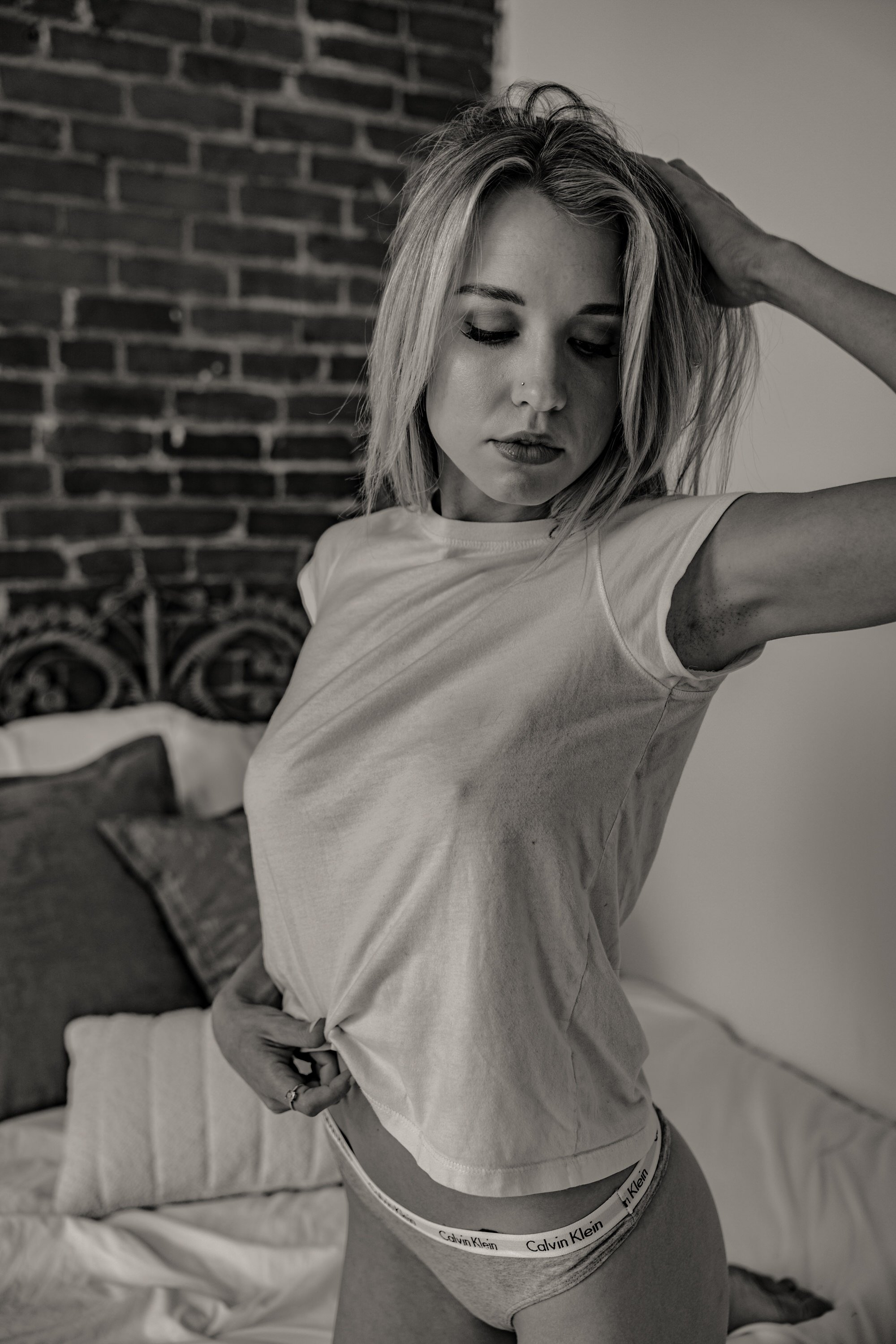 A woman with blonde hair and a nose piercing, wearing a white t-shirt and Calvin Klein underwear, poses indoors against a brick wall and a white wall, with her hand resting on her head and looking downward.