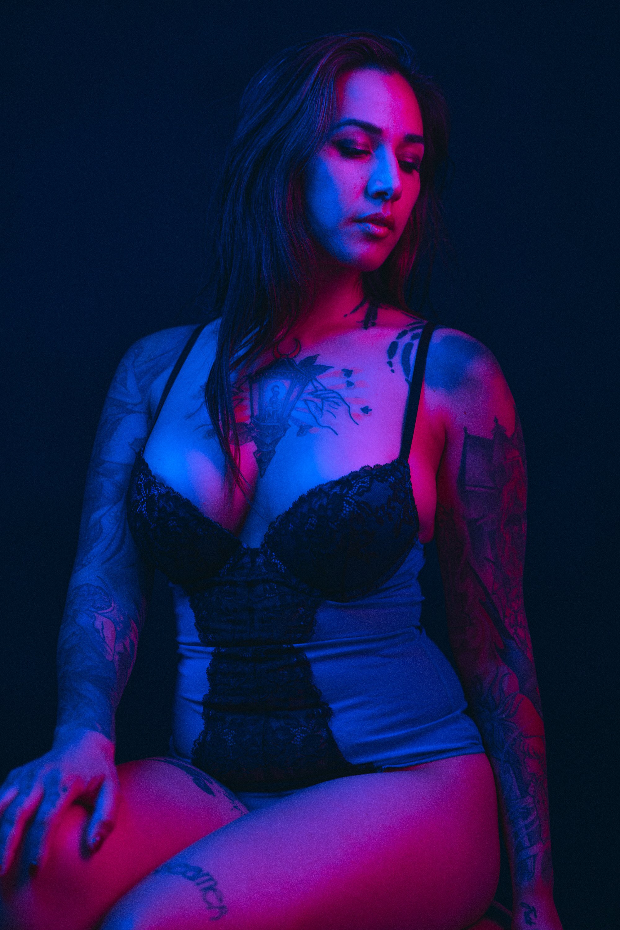 A woman with long dark hair and tattoos wearing a black lace top and gray shorts, sitting in a dimly lit space illuminated by purple and pink neon lights.