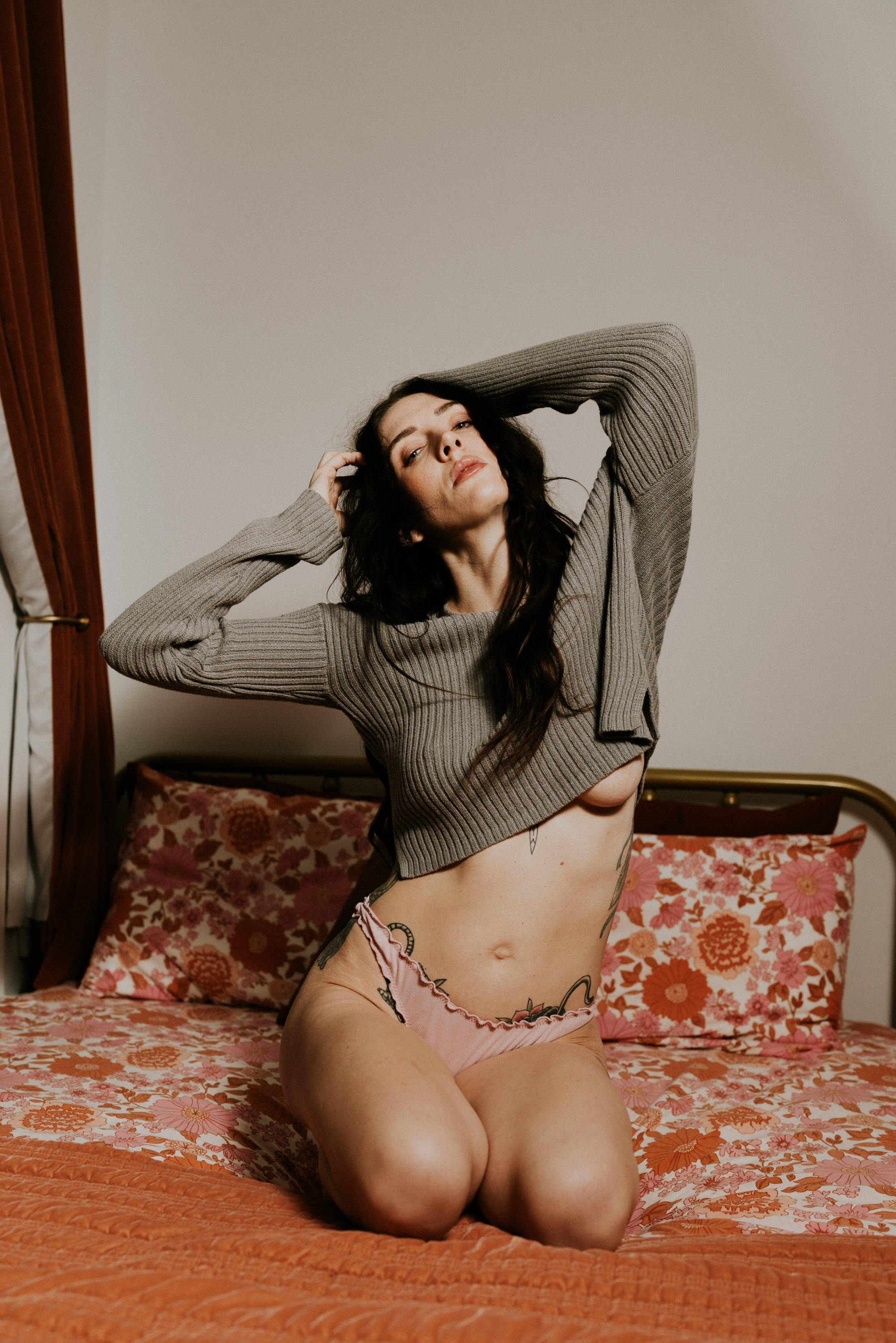 A woman with long dark hair wearing a gray knit sweater and pink underwear with a floral pattern, kneeling on a bed with a floral bedspread, posing with her hands behind her head.