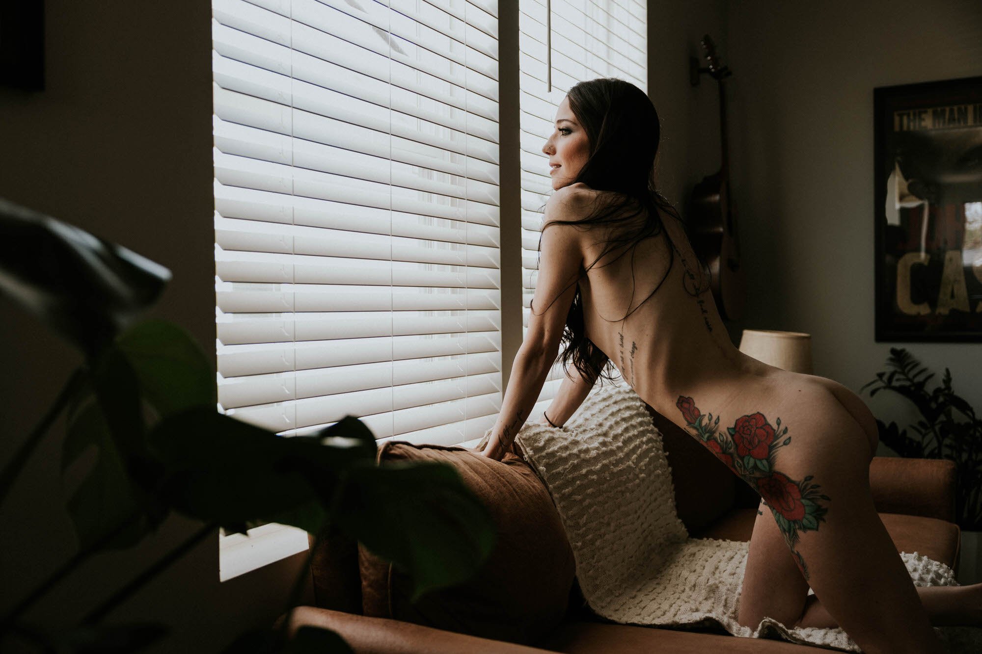 A woman with tattoos on her arm and back sitting on a bed by a window with white blinds, looking out, in a dimly lit room.