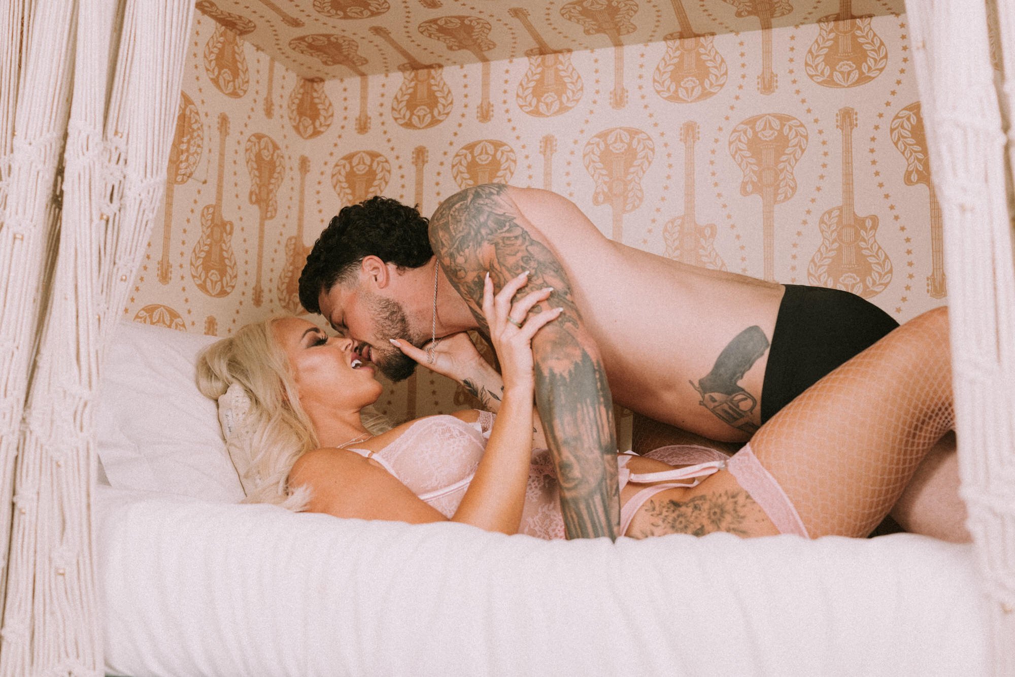 A man with tattoos and a woman with blonde hair lying in a bed, smiling and touching each other's faces.