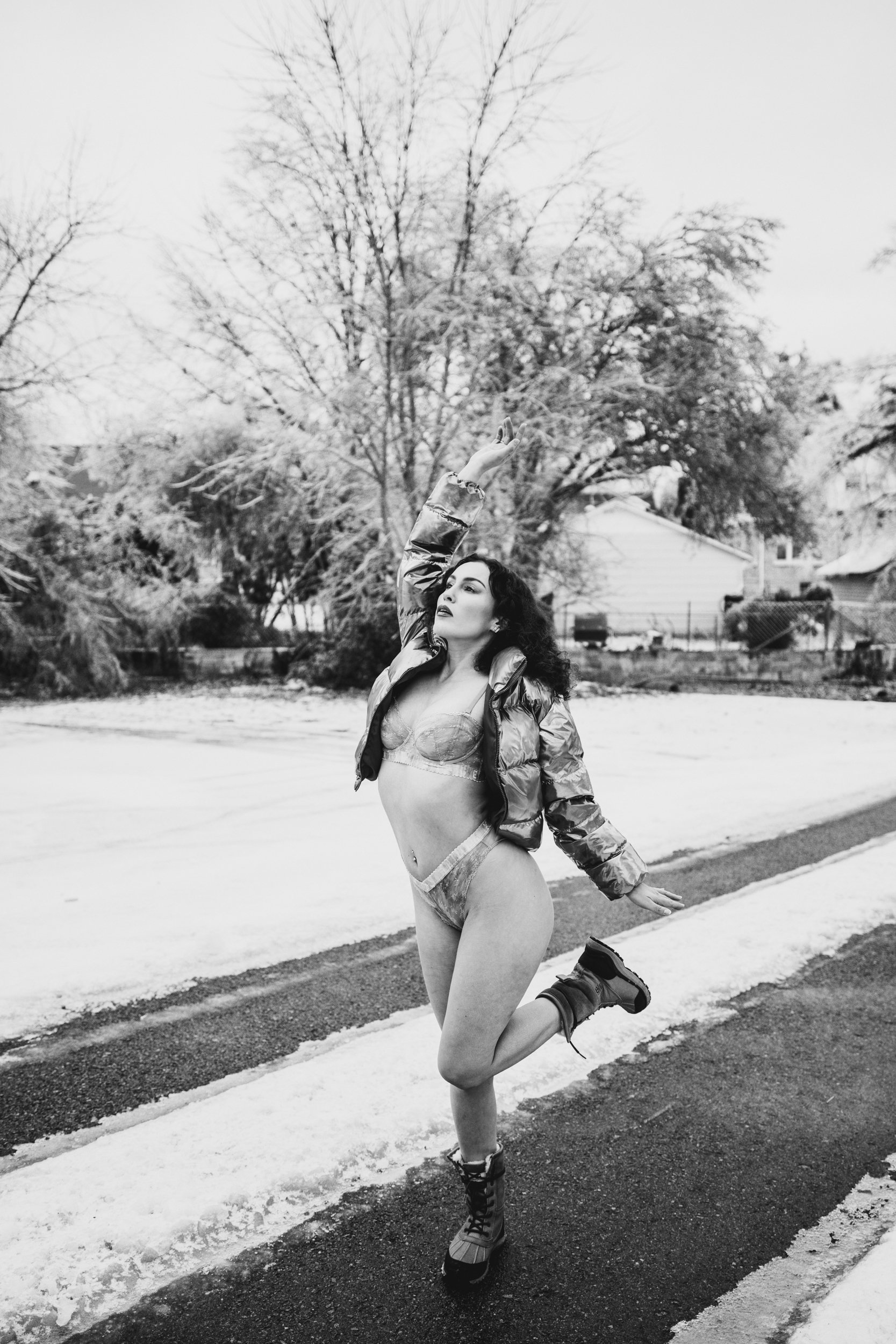 A woman in underwear, a puffy jacket, and winter boots is skating on a street covered with snow and ice. She is mid-stride with one leg in the air and arms raised, outdoors in a neighborhood with trees and houses in the background.