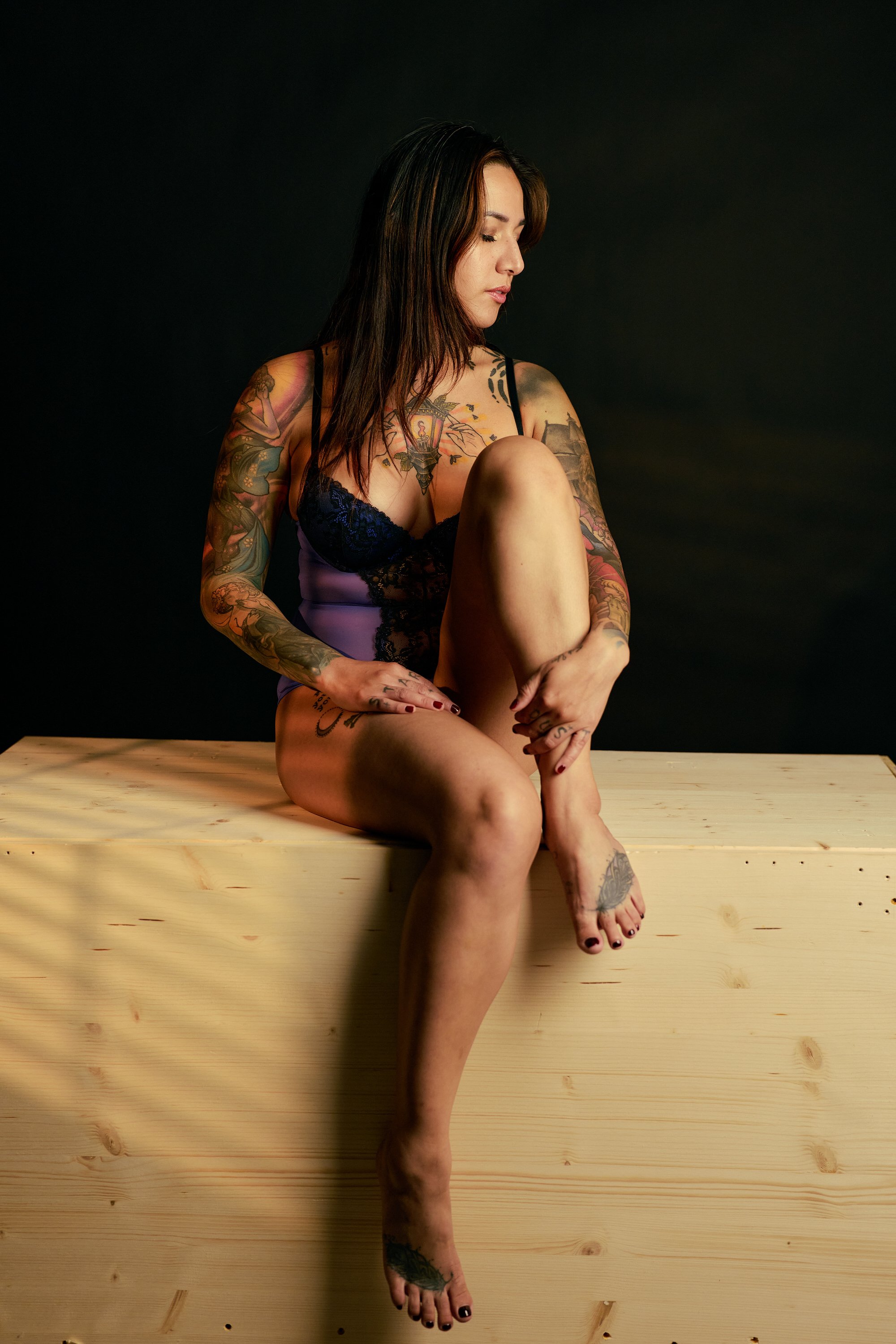 A tattooed woman seated on a wooden bench with dark background, wearing purple lace lingerie, with her eyes closed and looking down, with one knee up, displaying multiple tattoos on her arms, chest, and legs.