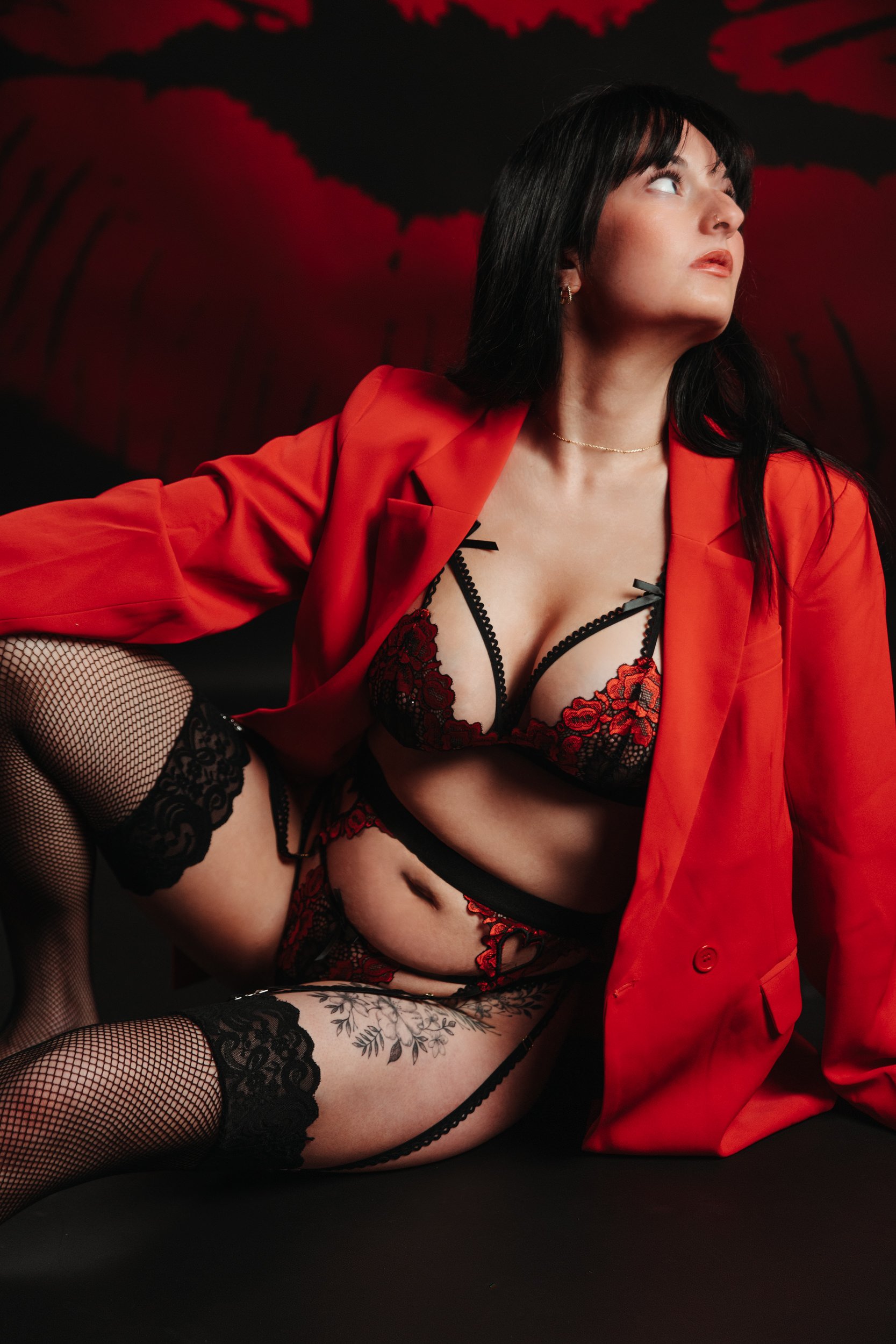 Woman in black and red lingerie, fishnet stockings, partially covered by a red blazer, posing against a dark background with red and black abstract art.