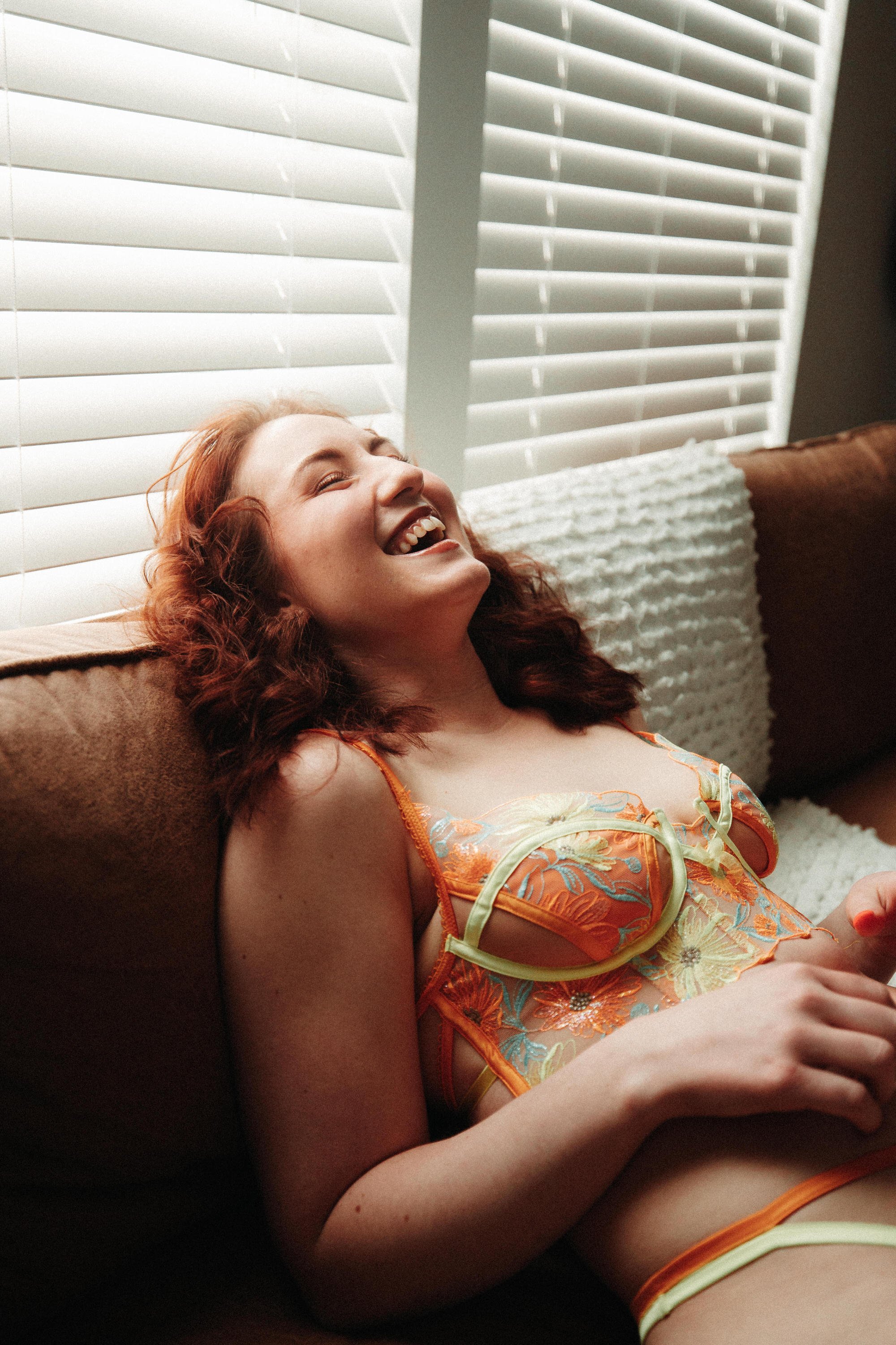 A woman with red hair and a big smile is sitting on a couch near a window with white blinds. She is wearing an orange floral lingerie top and appears to be relaxed and happy, possibly laughing.