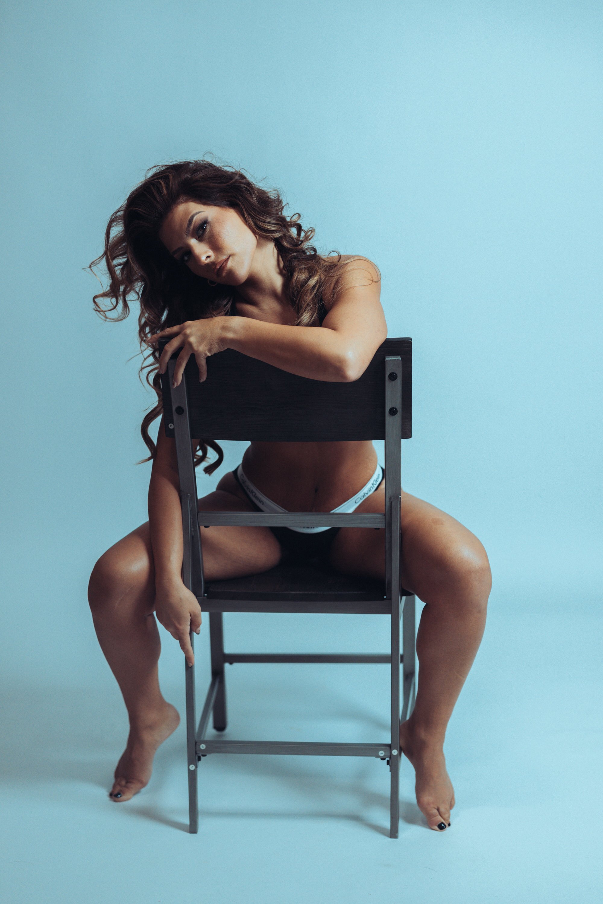 A woman with long, curly hair poses with a black chair and a metal step stool, sitting on the floor wearing Calvin Klein underwear, against a plain light blue background.