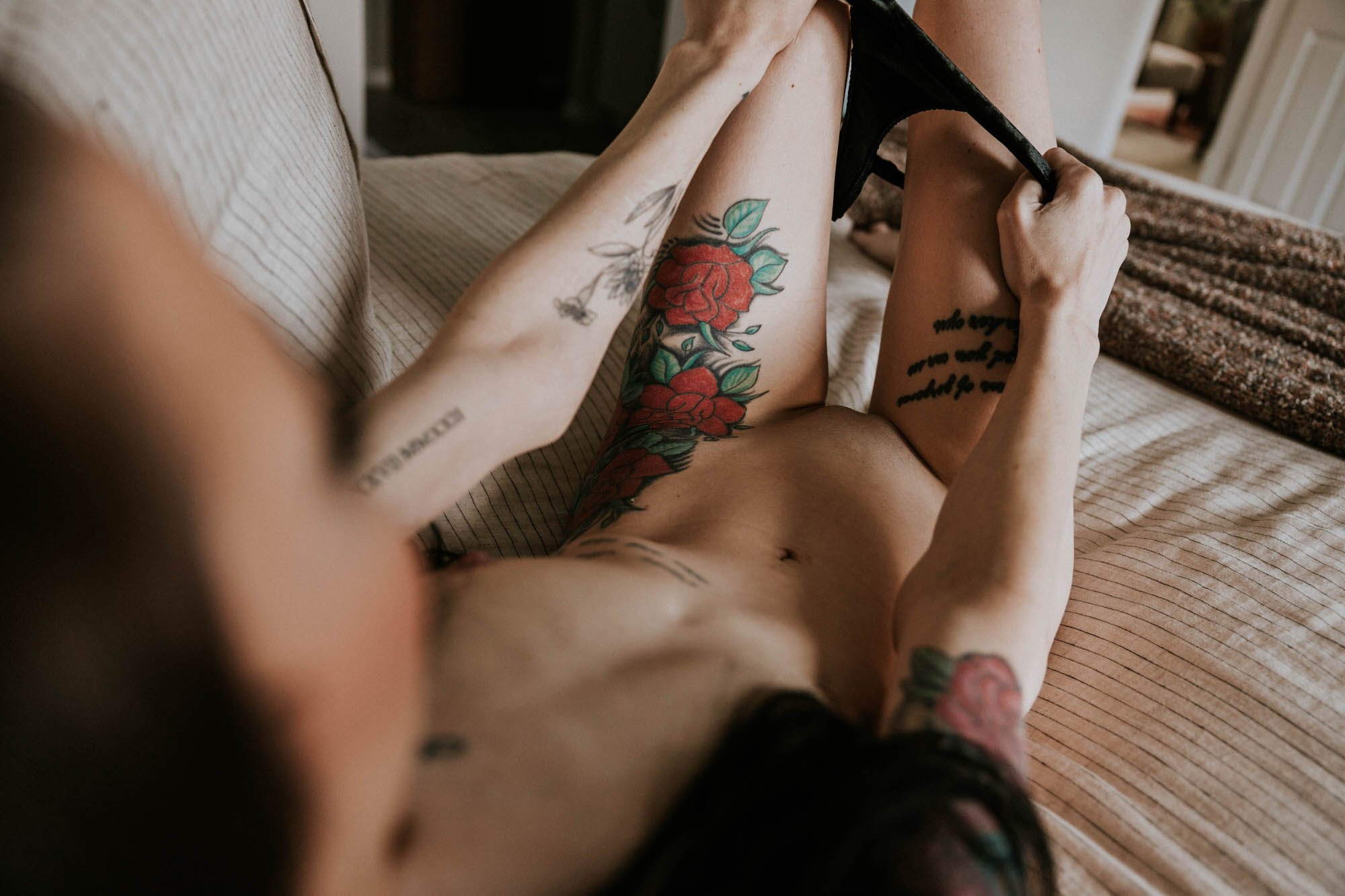 A person with colorful tattoos on their arm and leg is lying on a bed, holding a black high heel shoe.