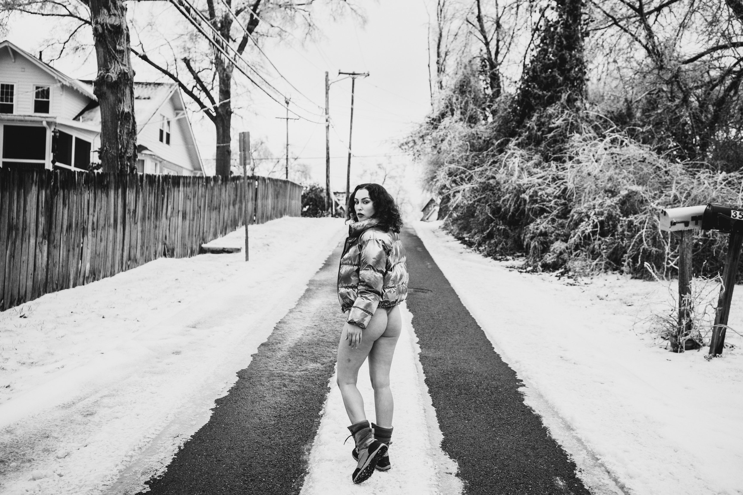 Black and white photo of a woman standing on a snowy road, wearing a puffer jacket, boots, and ski goggles, with trees, houses, and utility poles in the background.