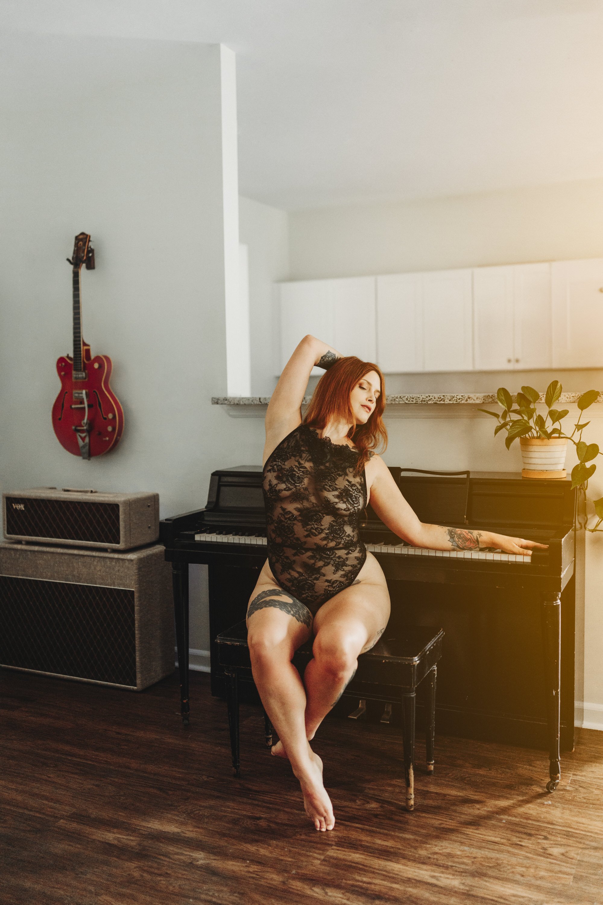 A woman with red hair in black lace lingerie sitting on a piano bench, with tattoos on her thigh and arm, her eyes closed, one arm raised behind her head, in a room with a guitar hanging on the wall and an amplifier, a potted plant on a piano, and wo